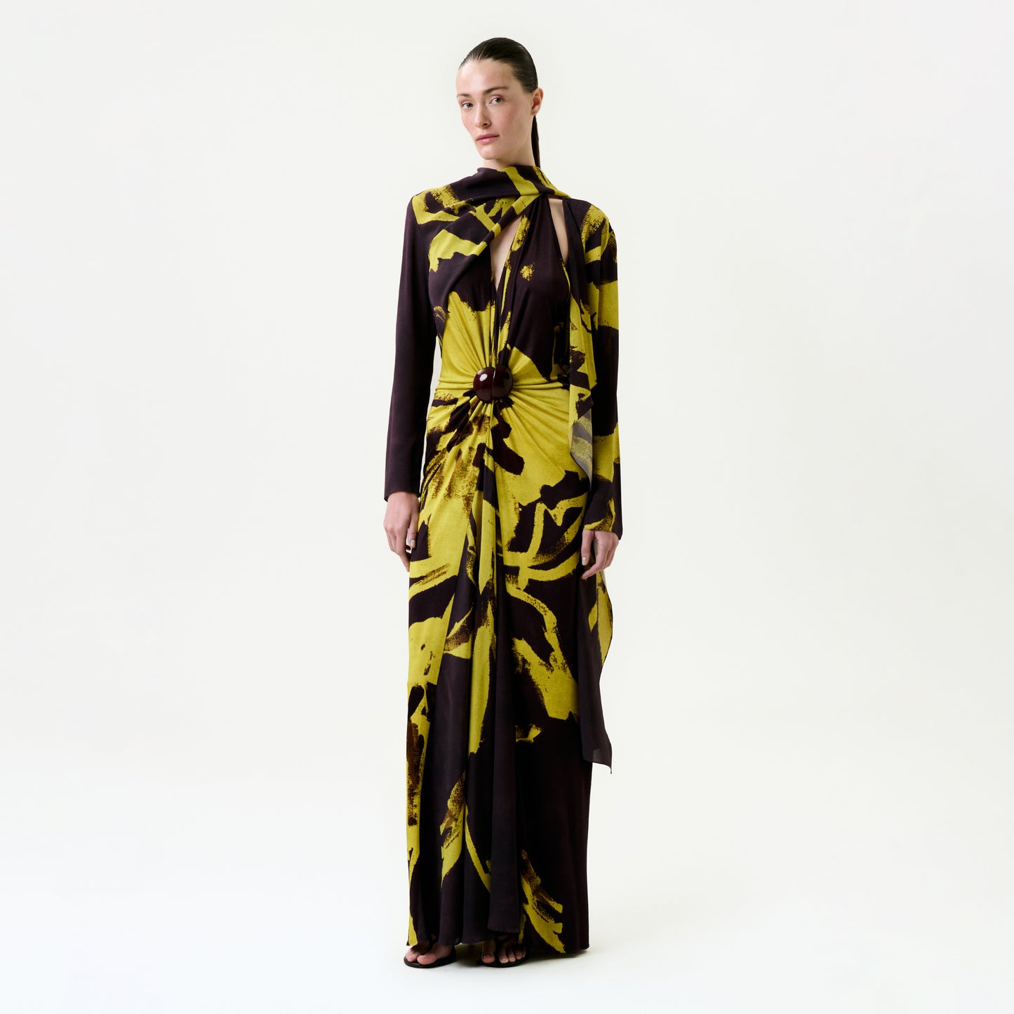 A woman stands against a white background wearing the Luisanna Cape, a long-sleeved, floor-length dress with a bold yellow and brown abstract floral print, crafted from luxurious Italian fabric and featuring a cape-like scarf at the neckline.