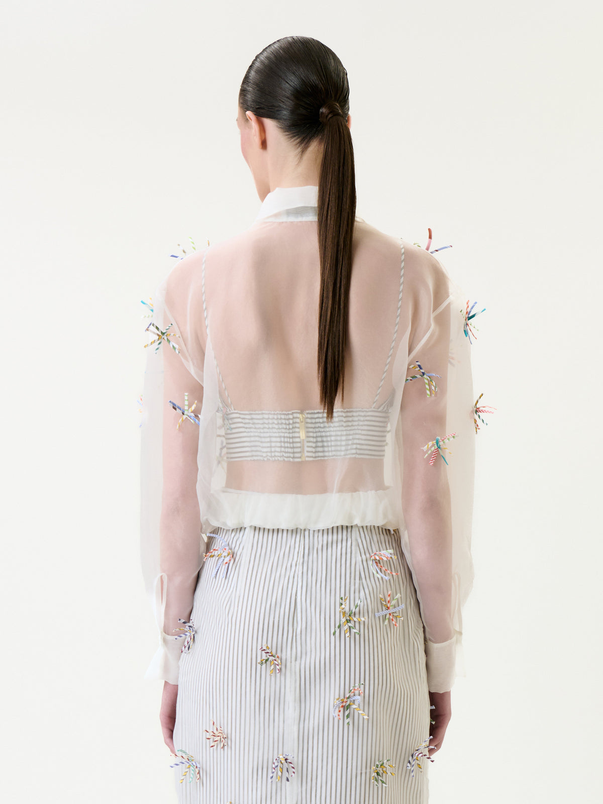 Seen from behind against a plain light background, a woman with straight dark hair in a low ponytail models the Katniss Jacket—a sheer white top adorned with colorful embellishments—paired with a striped skirt.