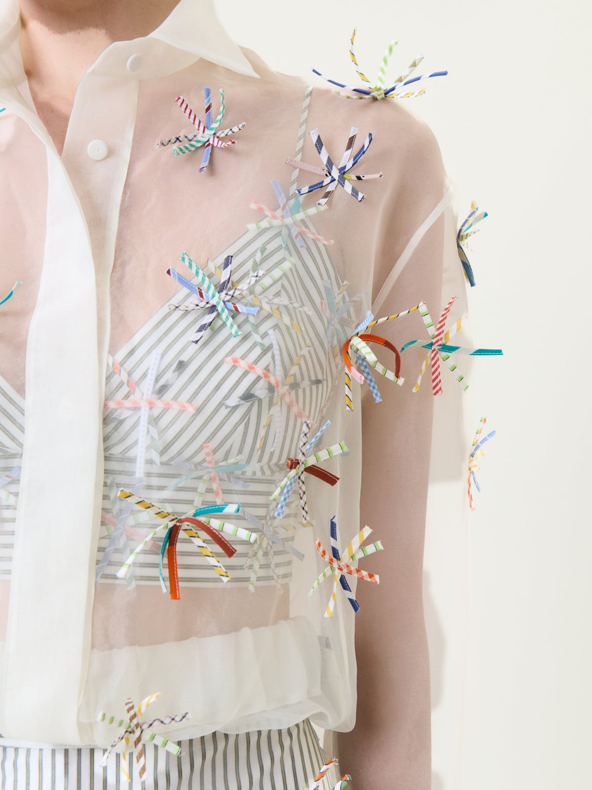 The Katniss Jacket is a sheer white jacket featuring vibrant ribbon-like embellishments bursting across the fabric, worn over a white-striped top. Only the torso is shown.