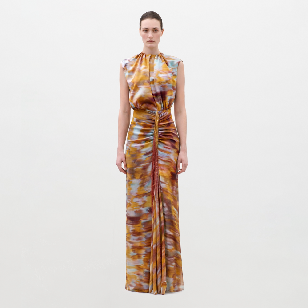 A woman faces forward in the Winona Dress, a sleeveless, floor-length gown with a ruched front and an abstract pattern in orange, yellow, blue, and brown, set against a plain white background.