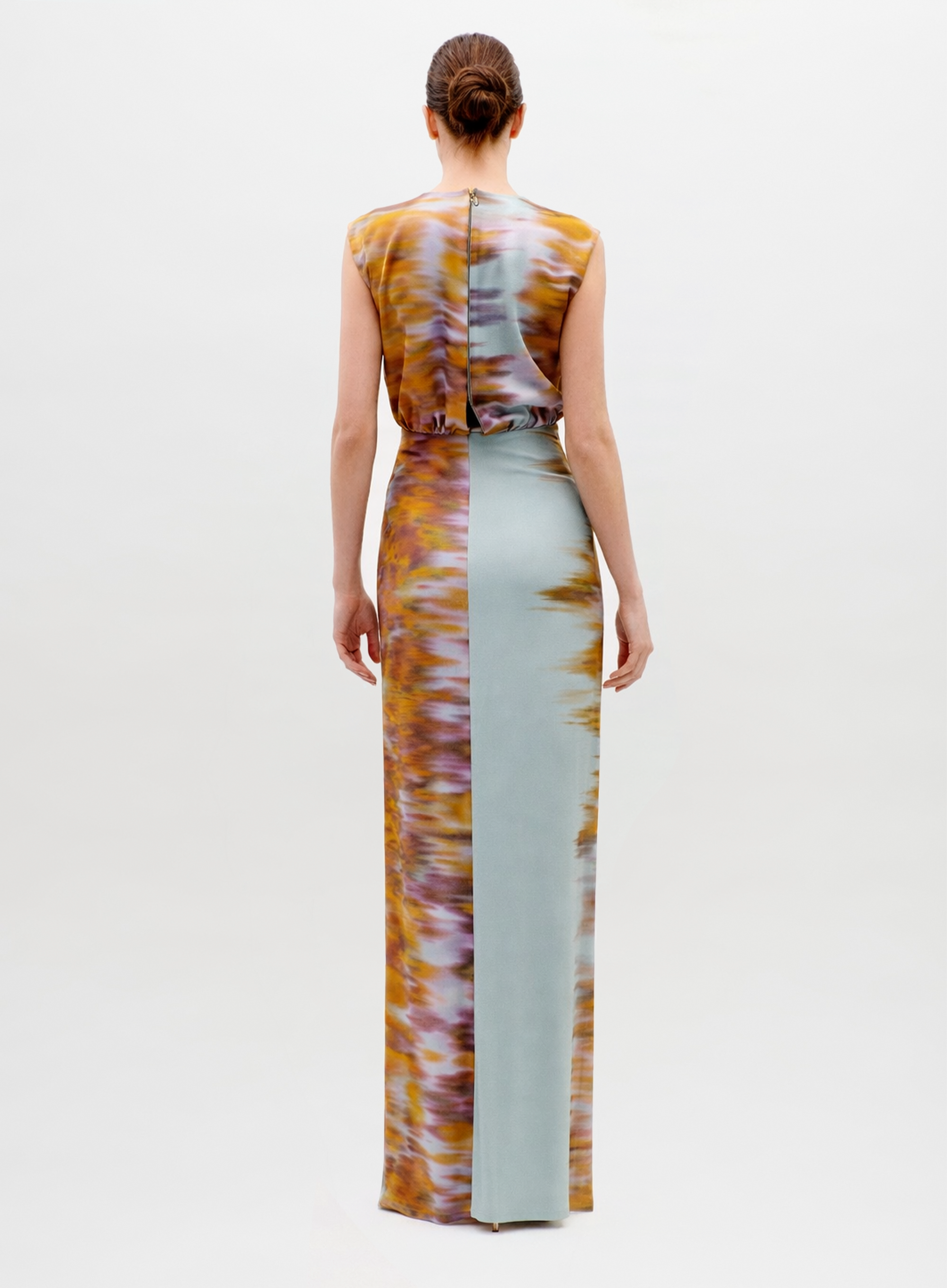 A woman with a neat low bun faces away, wearing the sleeveless, floor-length Winona Dress featuring a split design: one side in light blue and the other in a brown and yellow textured pattern.