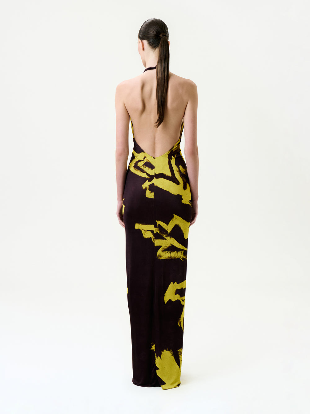 A person with long, straight hair in a low ponytail wears the Aiko Dress—a backless, floor-length maxi made in Colombia with a dark base and bold yellow abstract patterns—standing against a plain white background.