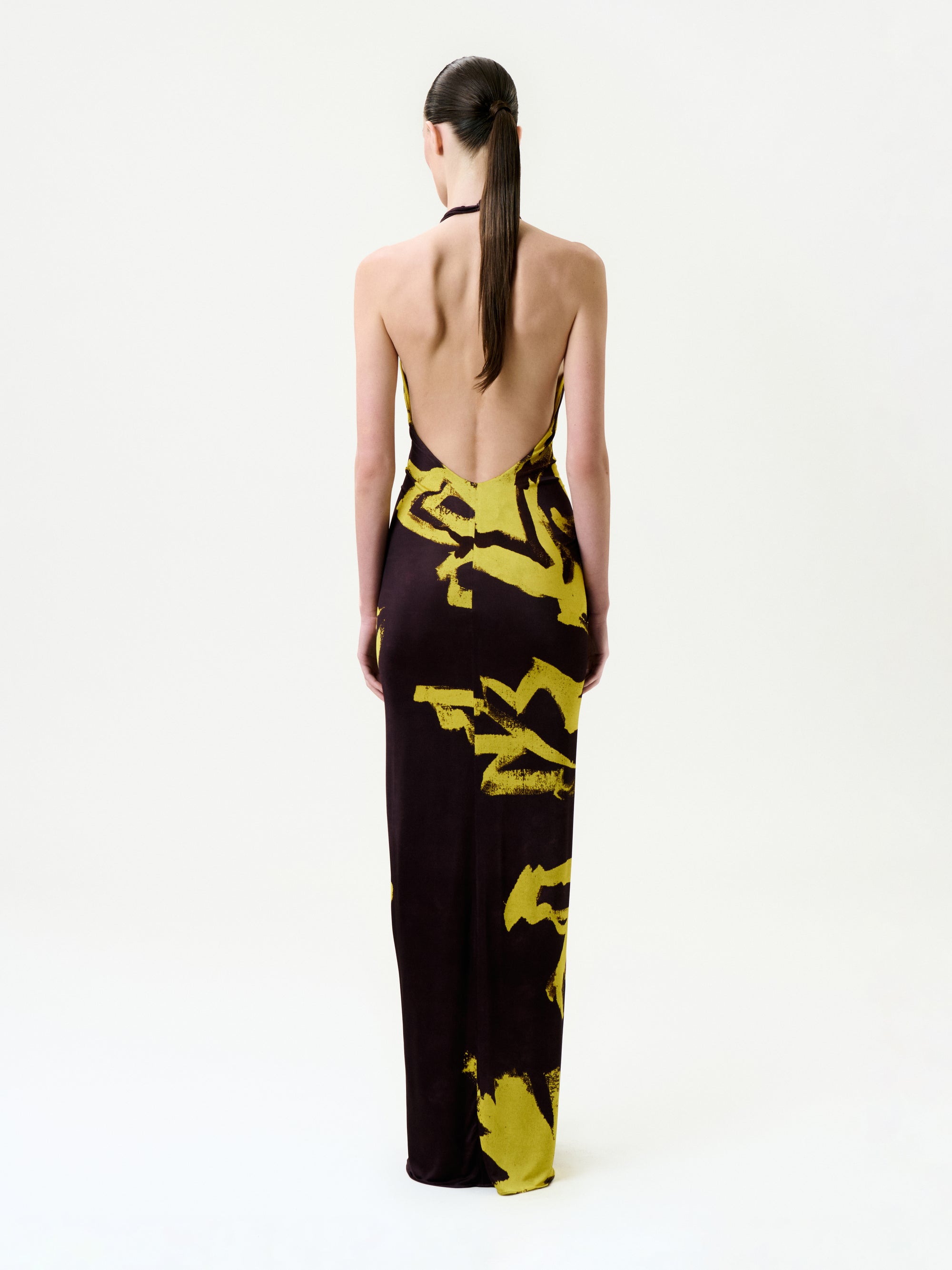 A person with long, straight hair in a low ponytail wears the Aiko Dress—a backless, floor-length maxi made in Colombia with a dark base and bold yellow abstract patterns—standing against a plain white background.