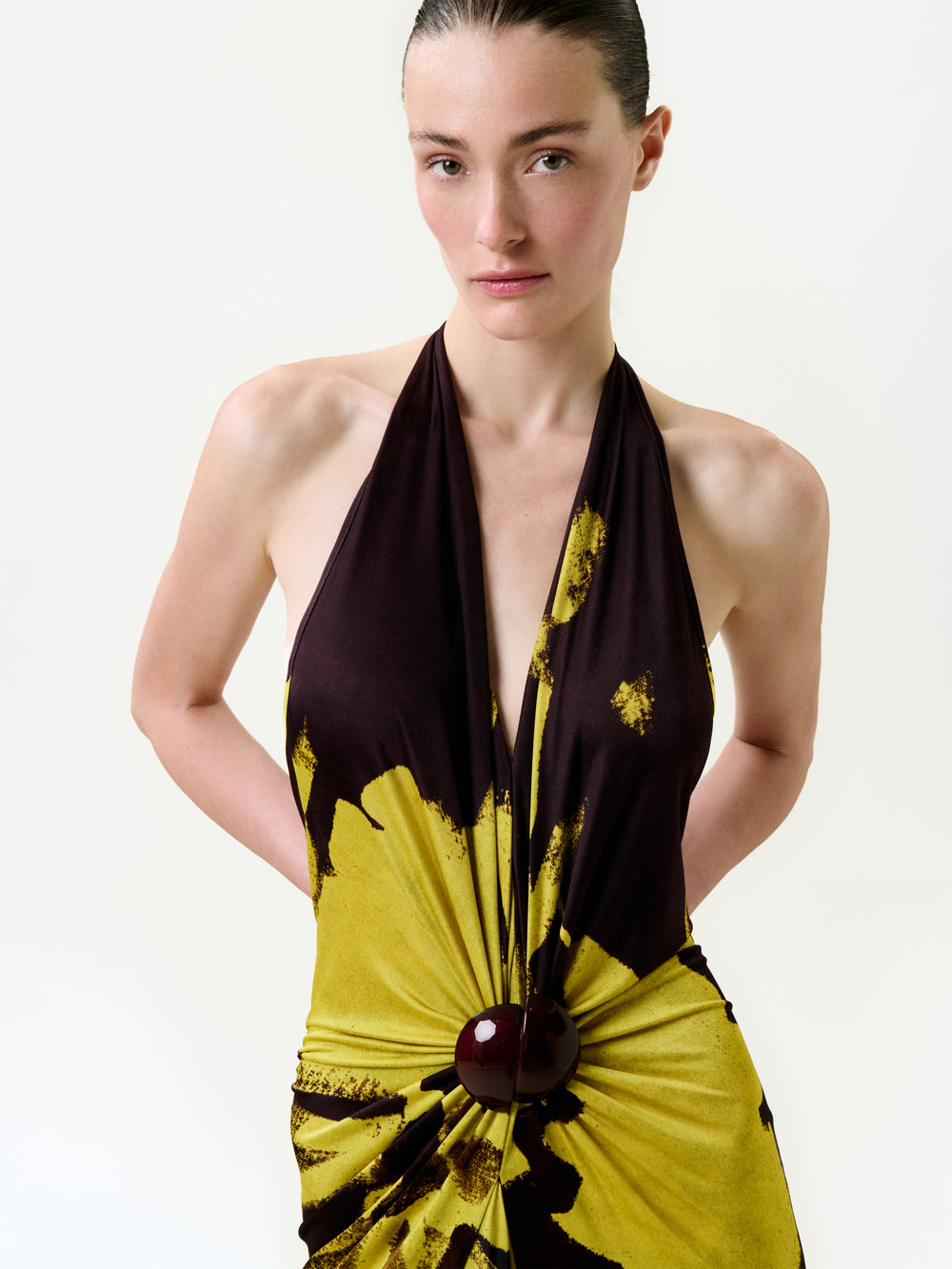 A woman with fair skin and dark hair pulled back wears the Aiko Dress, a sleeveless yellow and dark brown halter maxi with a deep neckline and round waist detail, made from Italian fabric, standing against a plain light background.