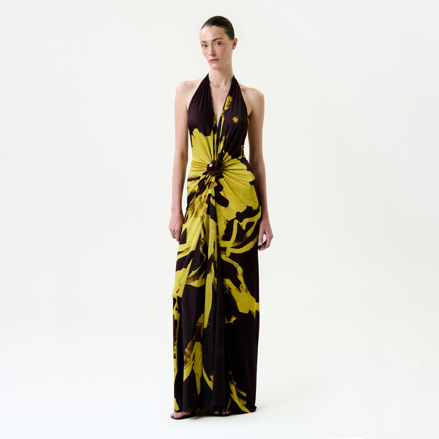 A woman stands against a white background wearing the Aiko Dress, a bold yellow and black floral maxi with a central waist knot, crafted from premium Italian fabric. Her hair is pulled back as she looks straight ahead.