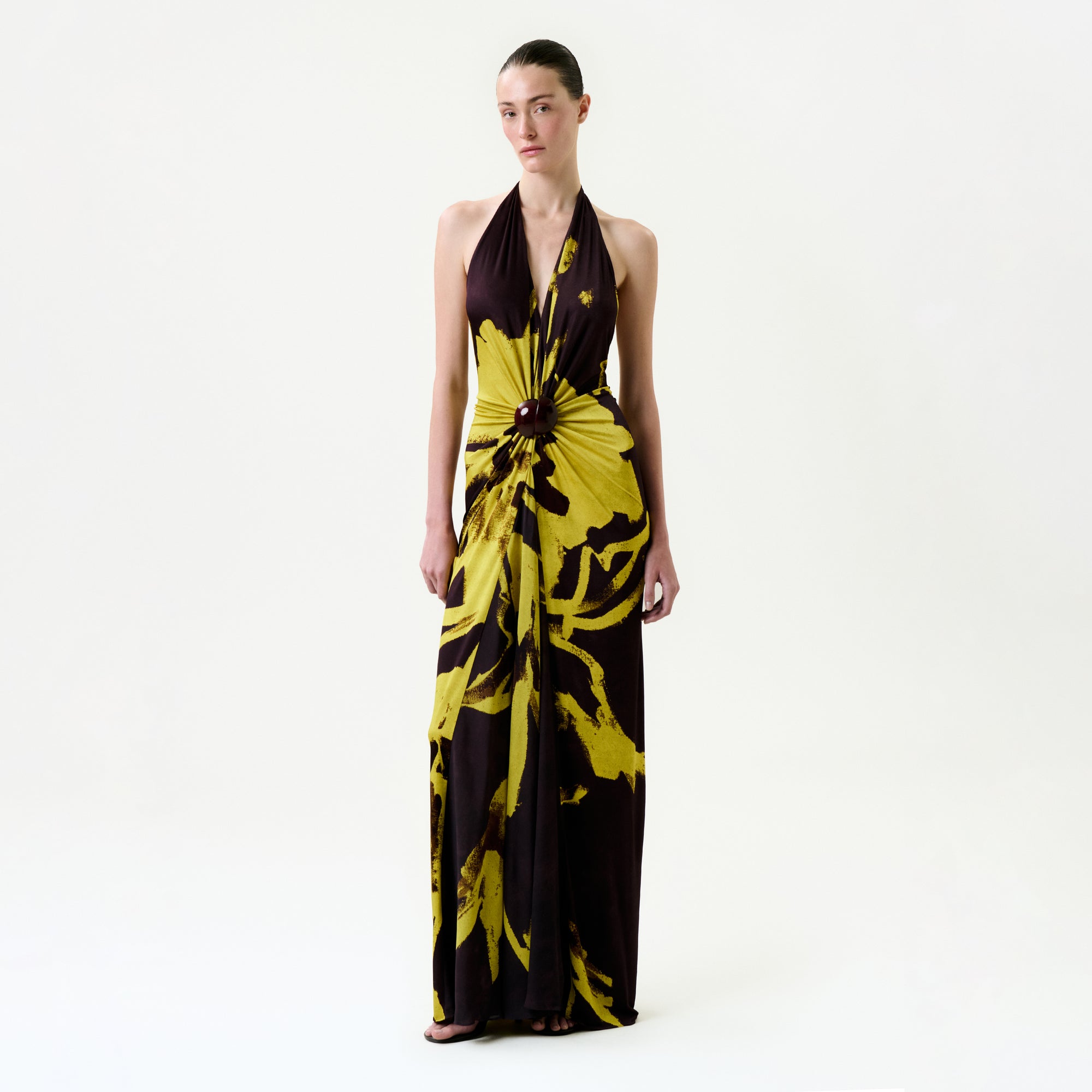 A woman stands against a white background wearing the Aiko Dress, a bold yellow and black floral maxi with a central waist knot, crafted from premium Italian fabric. Her hair is pulled back as she looks straight ahead.