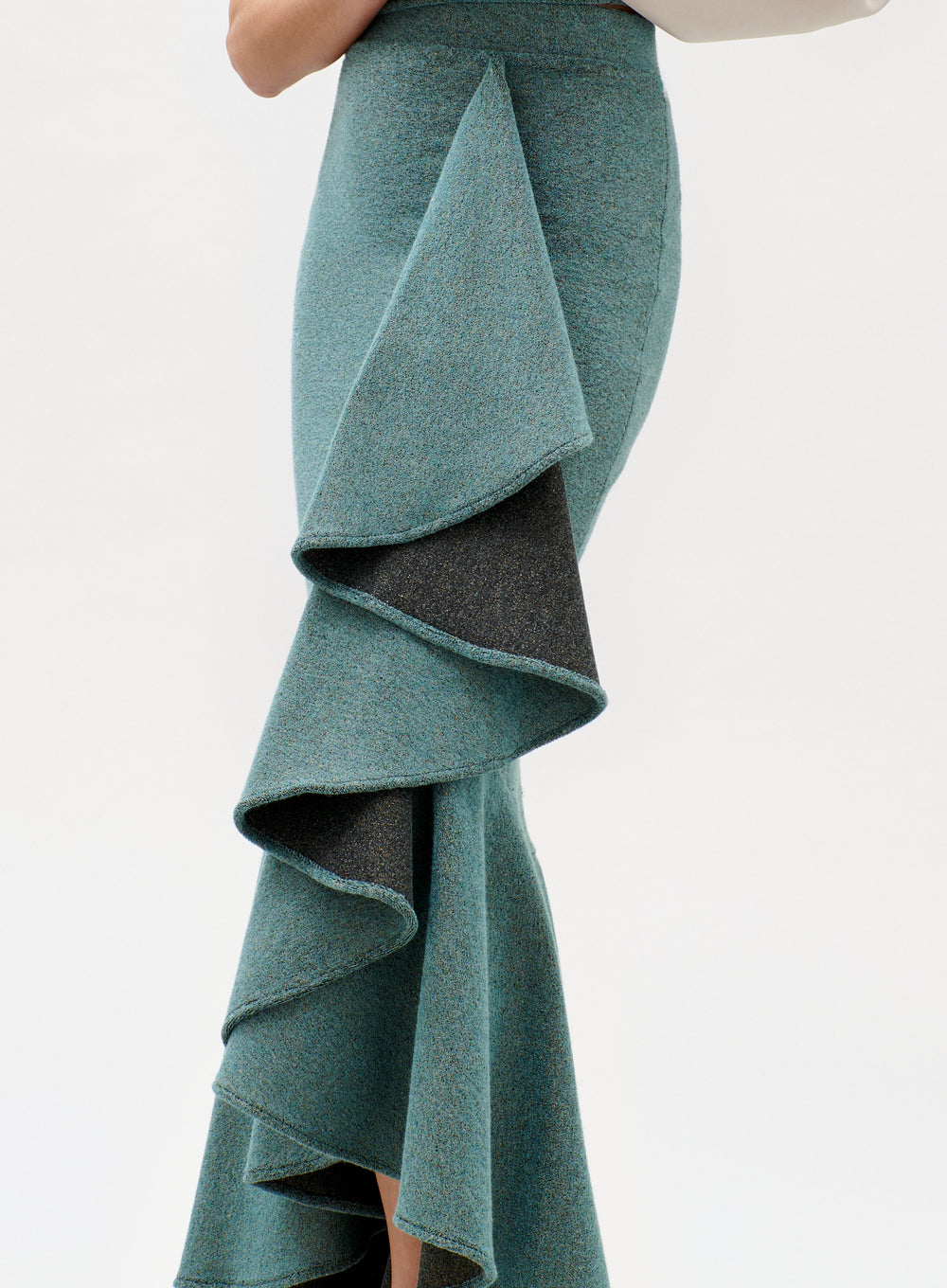 A person wears the Bambolera Skirt—a fitted, teal textured skirt featuring dramatic, asymmetrical layered ruffles—while standing against a plain white background.