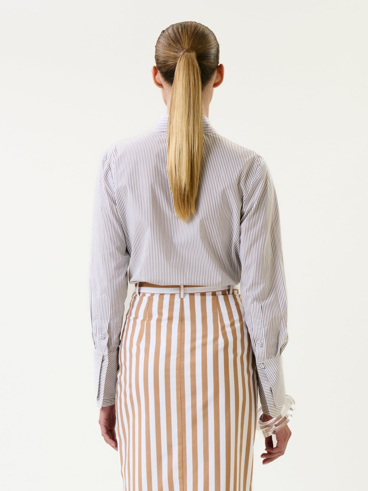 A person with long, straight blonde hair in a ponytail is seen from behind, wearing the Karena Shirt, which features thin stripes, paired with a skirt that has wide vertical beige and white stripes.