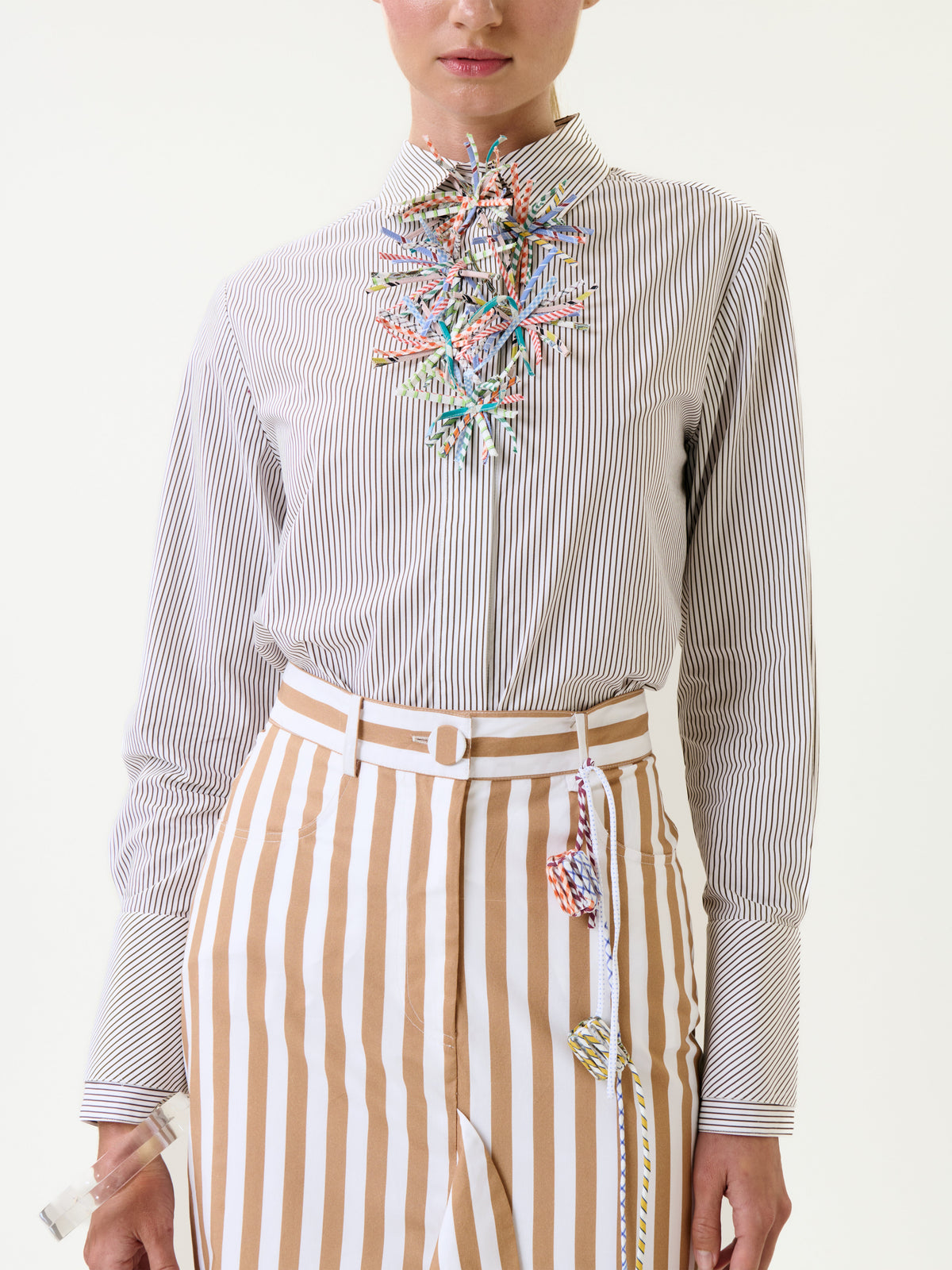 A person wearing the Karena Shirt—a white button-up shirt with thin black stripes—paired with tan-and-white striped high-waisted pants. Colorful decorative pieces hang from the collar and waistband. The background is plain white.