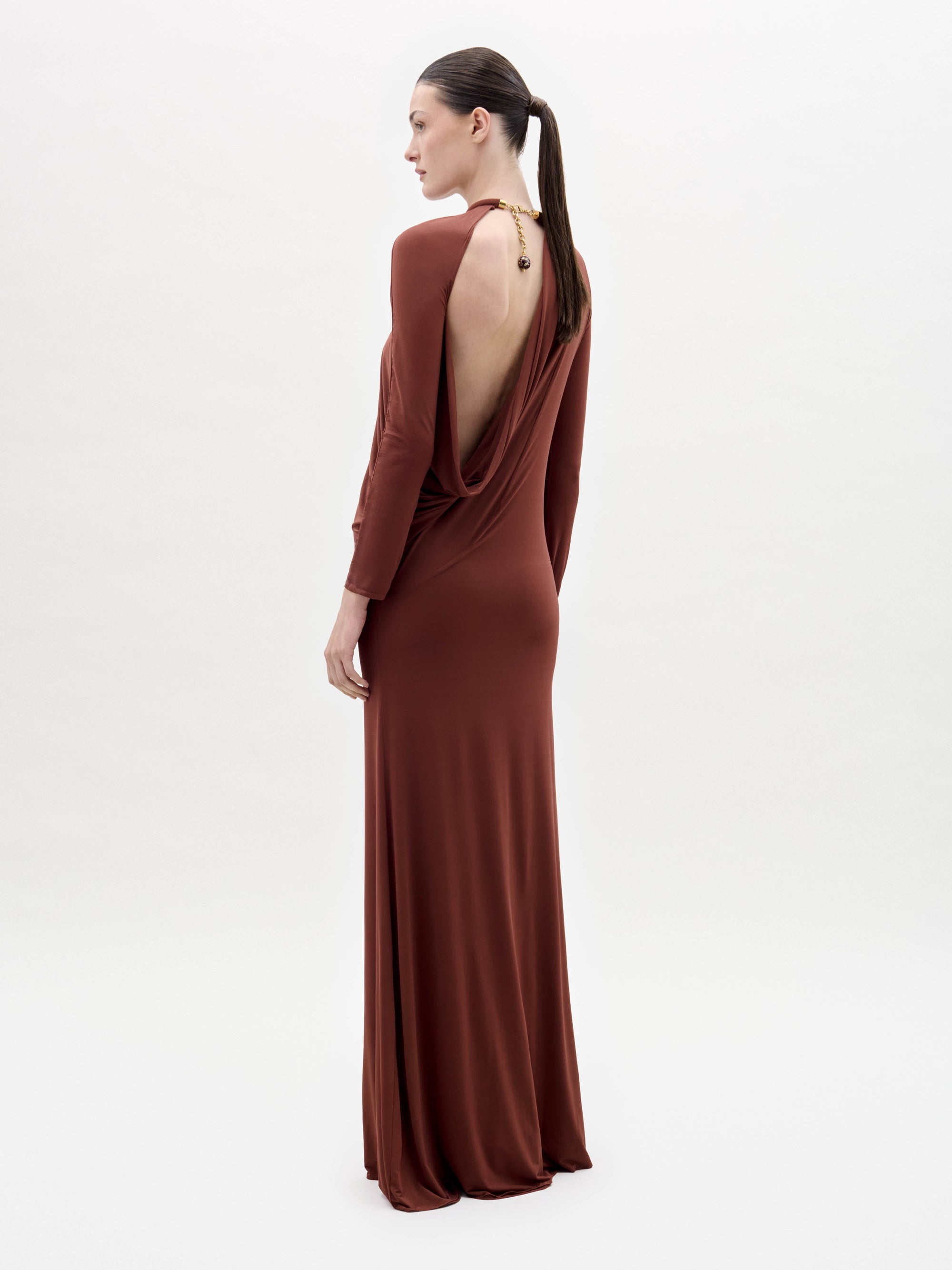 Someone stands against a plain white background wearing the Lais Dress, a long brown dress with long sleeves and a gathered neckline, styled with minimal accessories.