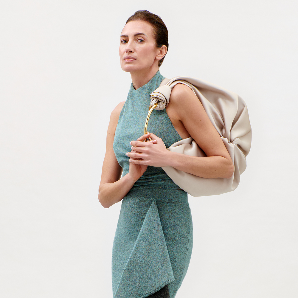 A woman wears the Lark Top in light teal, posing against a plain background with a large beige knotted bag featuring a gold handle on her shoulder, gazing confidently at the camera.