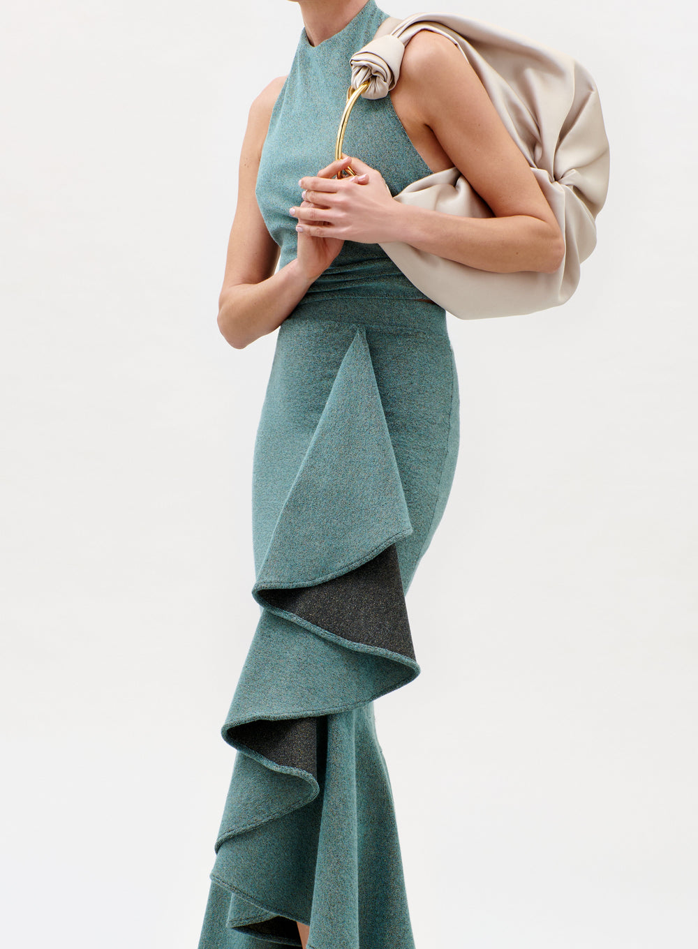 A woman models the Lark Top, a sleeveless teal piece with dramatic layered ruffles, paired with a large beige ruched handbag against a plain light background. Her head is cropped out of the image.