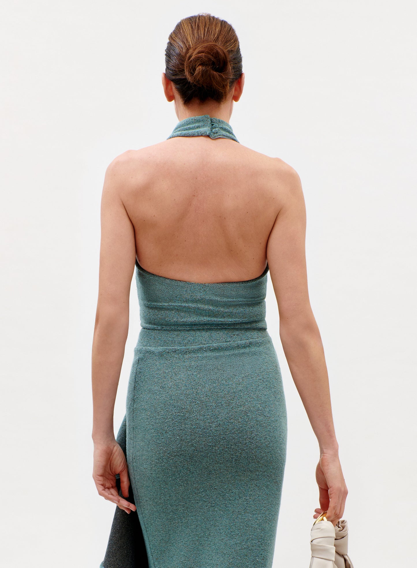 A woman with her hair in a neat bun models the sleeveless, teal Lark Top from the back, holding a light-colored handbag in her left hand against a plain, light background.