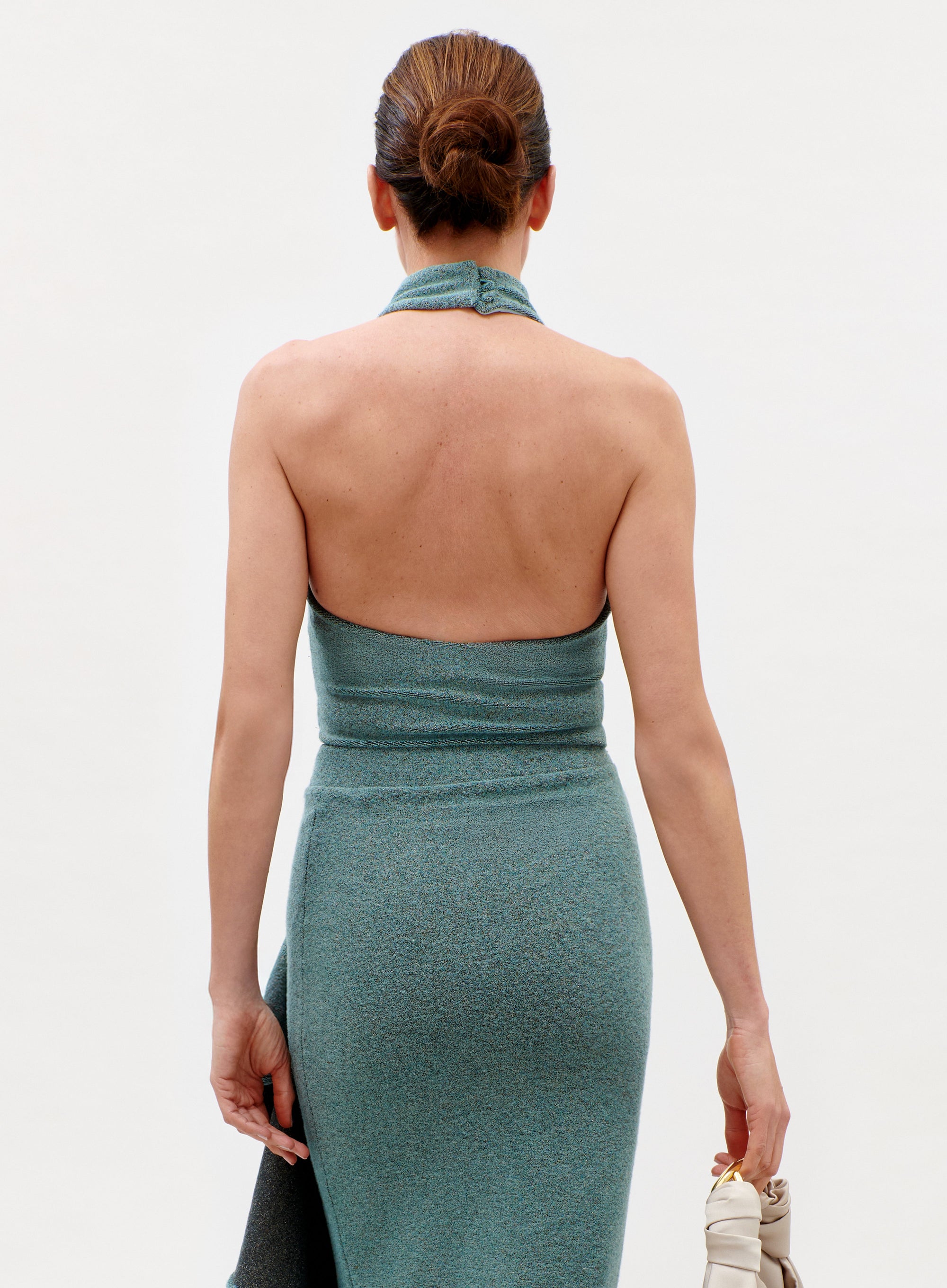 A woman with her hair in a neat bun models the sleeveless, teal Lark Top from the back, holding a light-colored handbag in her left hand against a plain, light background.