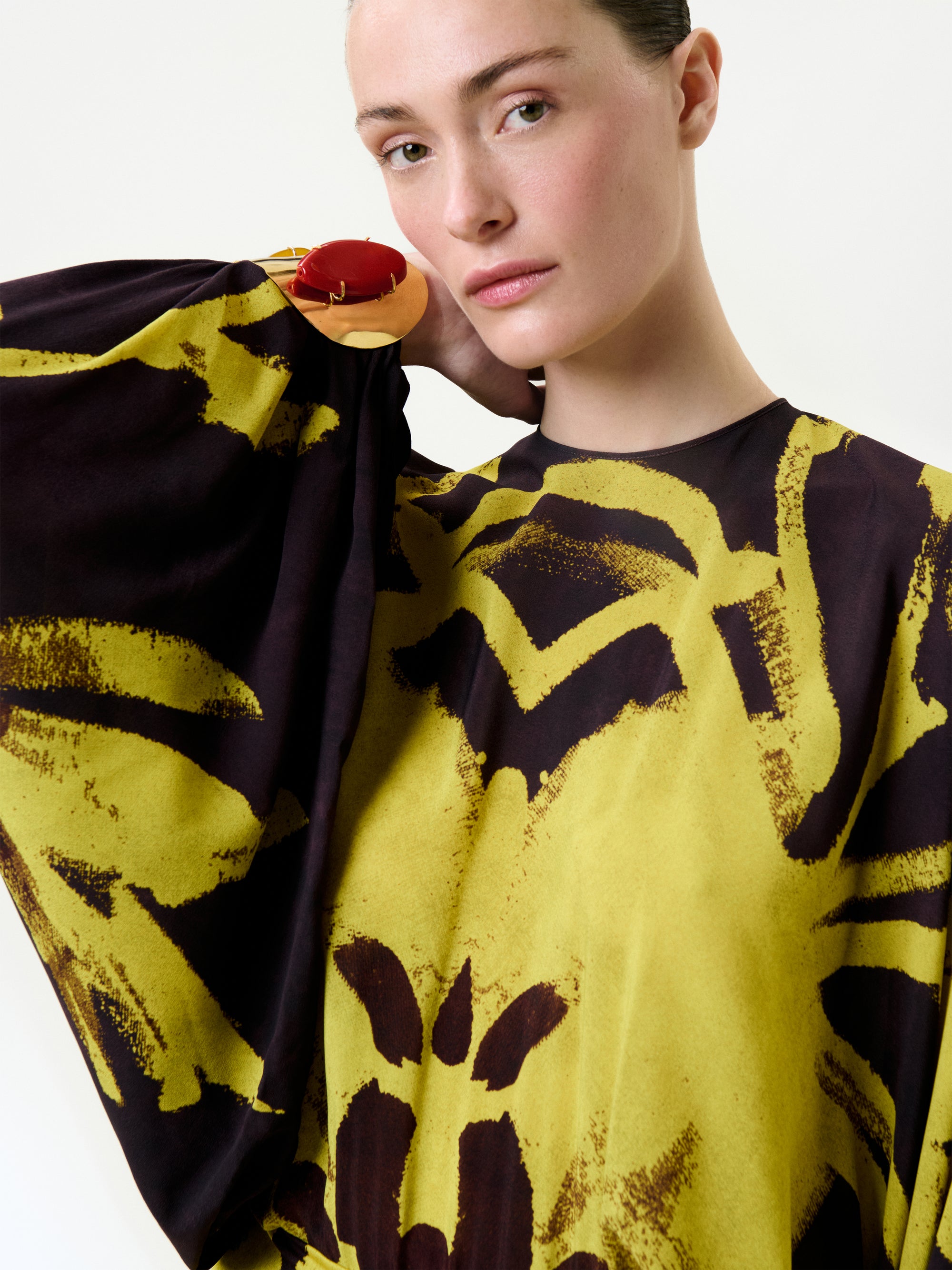 A woman with light skin and tied-back hair models the Kumiko Dress, a loose yellow and black patterned top made from Italian fabric with wide sleeves. She poses with one arm raised, displaying a red and gold bracelet against a plain backdrop.