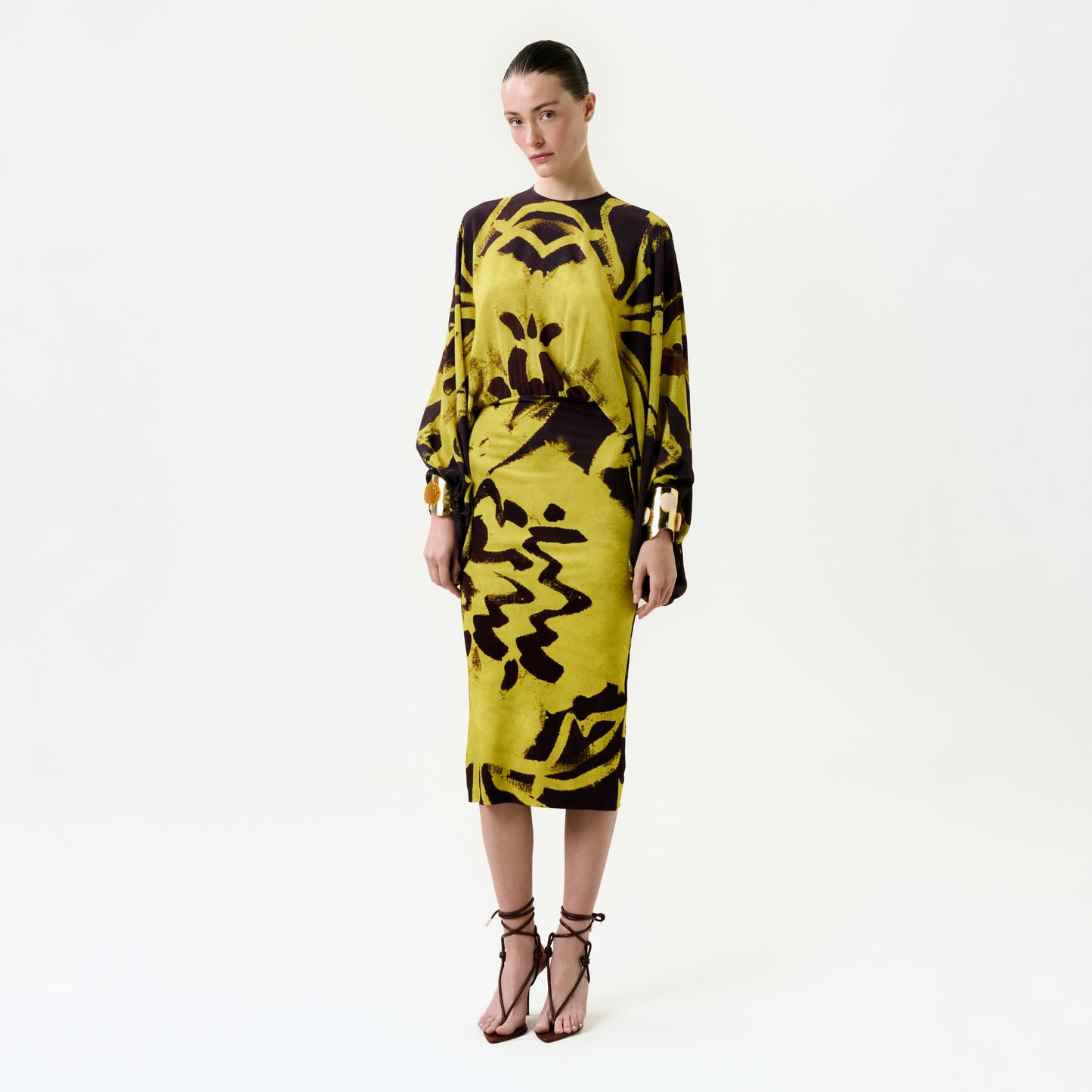 A woman stands against a plain white background, wearing the Kumiko Dress—a yellow and dark brown abstract-patterned midi dress—paired with brown strappy sandals and gold bracelets.