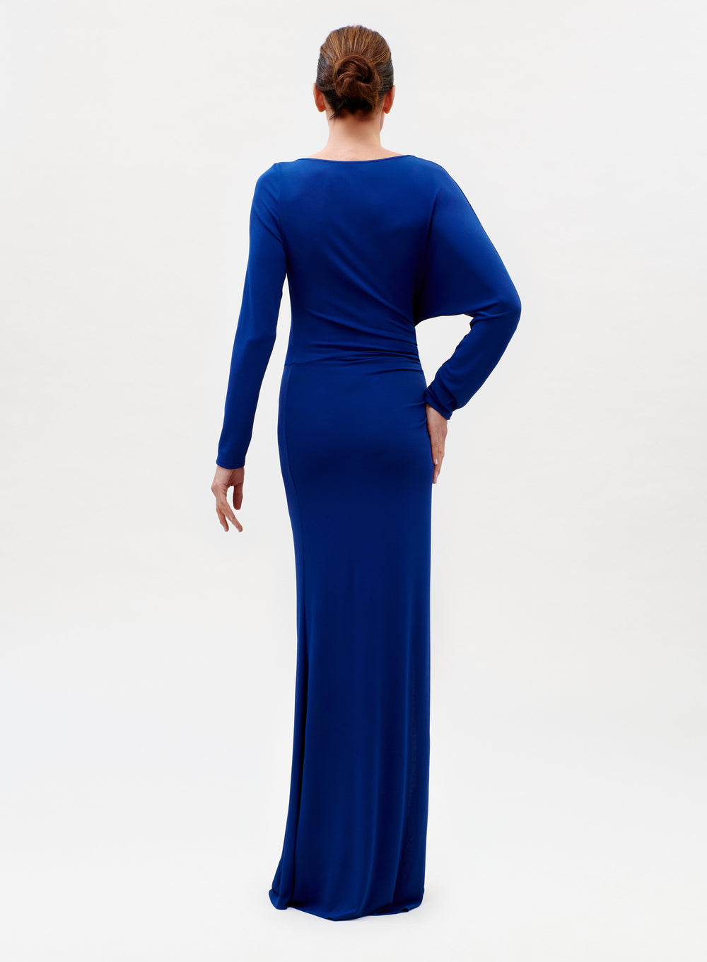 A woman with brown hair in a bun stands with her back to the camera, wearing the Laken Dress—a long-sleeved, floor-length royal blue gown—against a plain white background.