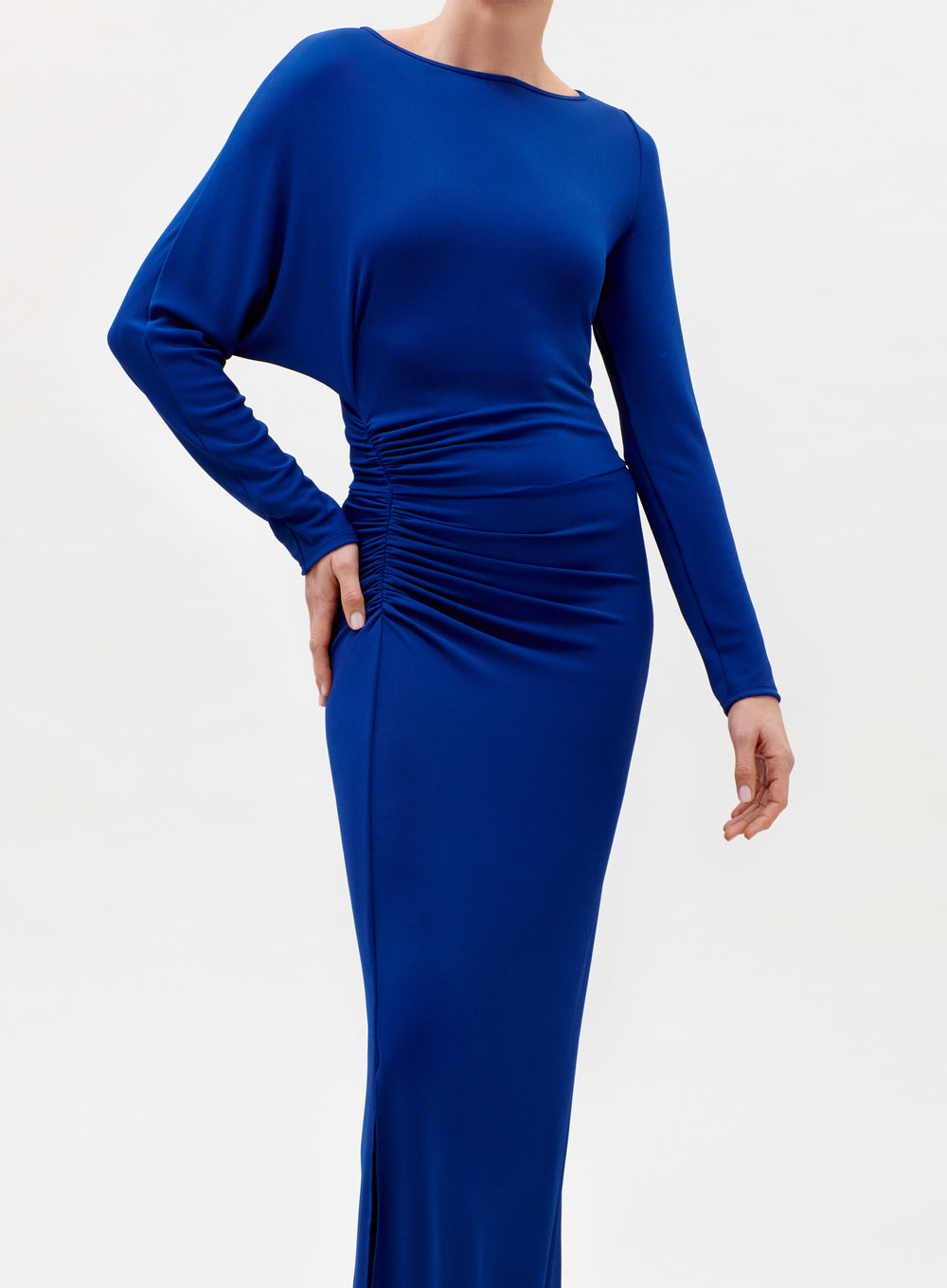 A person wearing the Laken Dress—a long-sleeved, royal blue, floor-length gown with side ruching—stands against a plain light background. The person's face is not visible.