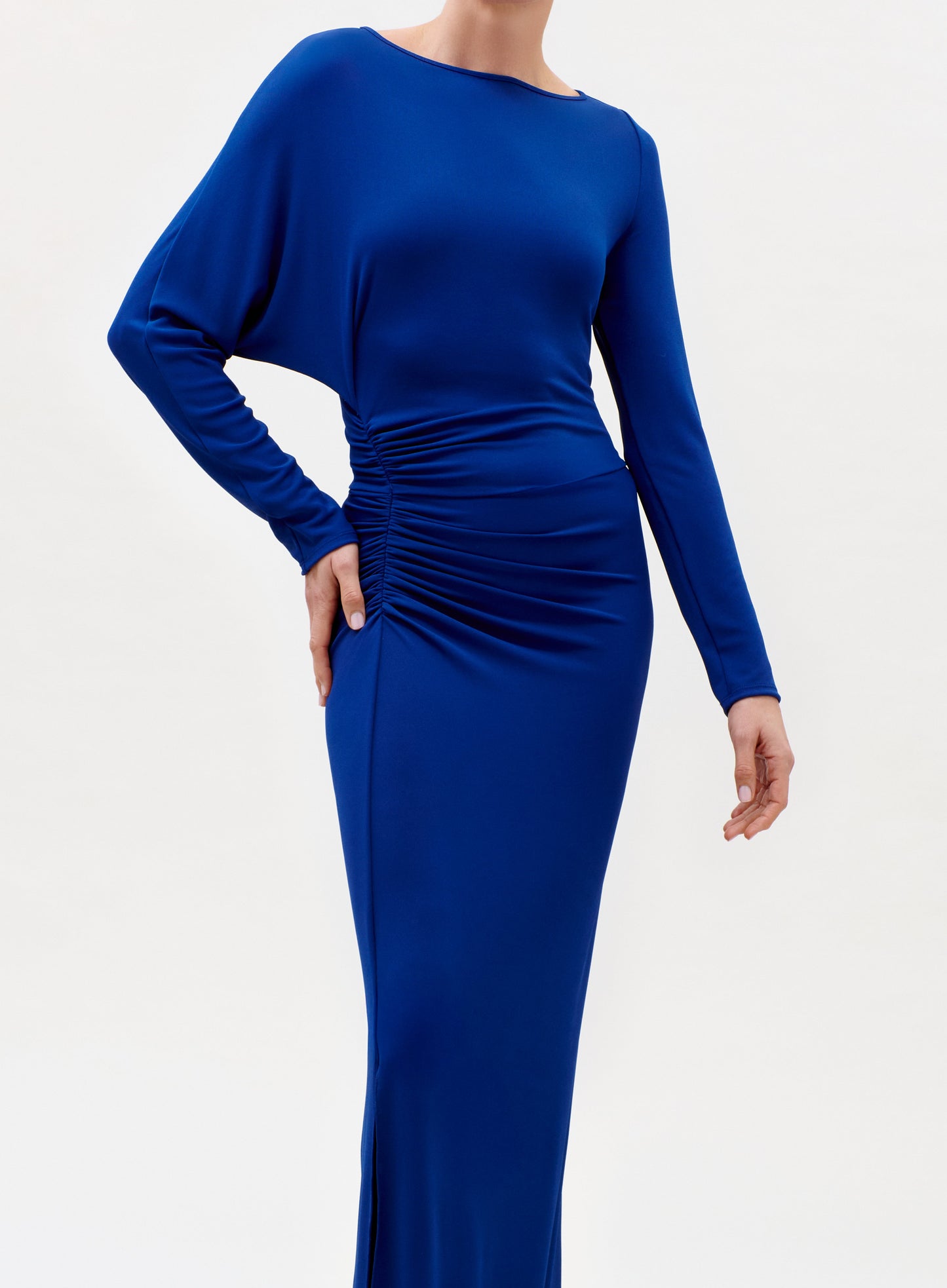 A person wearing the Laken Dress—a long-sleeved, royal blue, floor-length gown with side ruching—stands against a plain light background. The person's face is not visible.