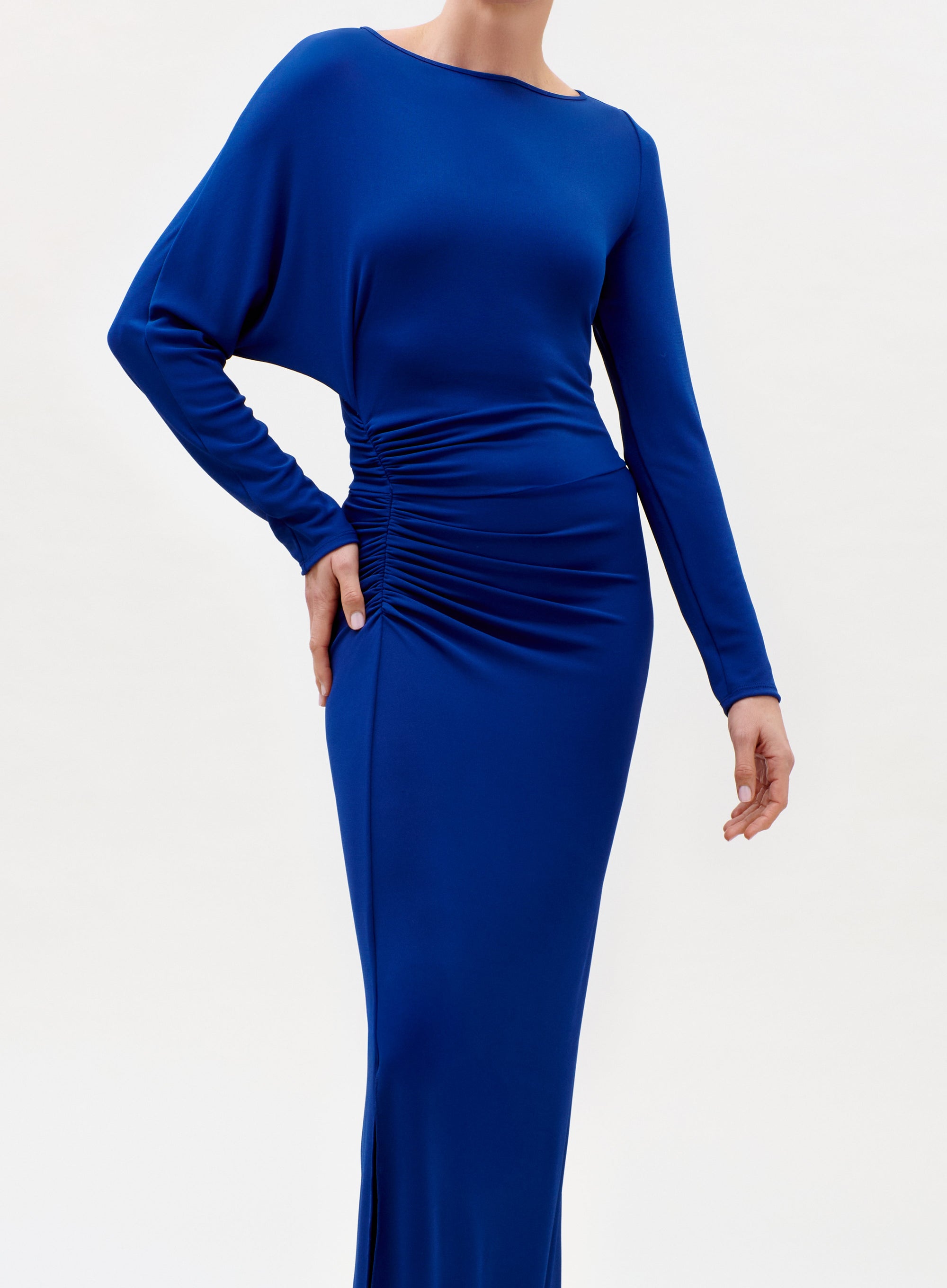 A person wearing the Laken Dress—a long-sleeved, royal blue, floor-length gown with side ruching—stands against a plain light background. The person's face is not visible.