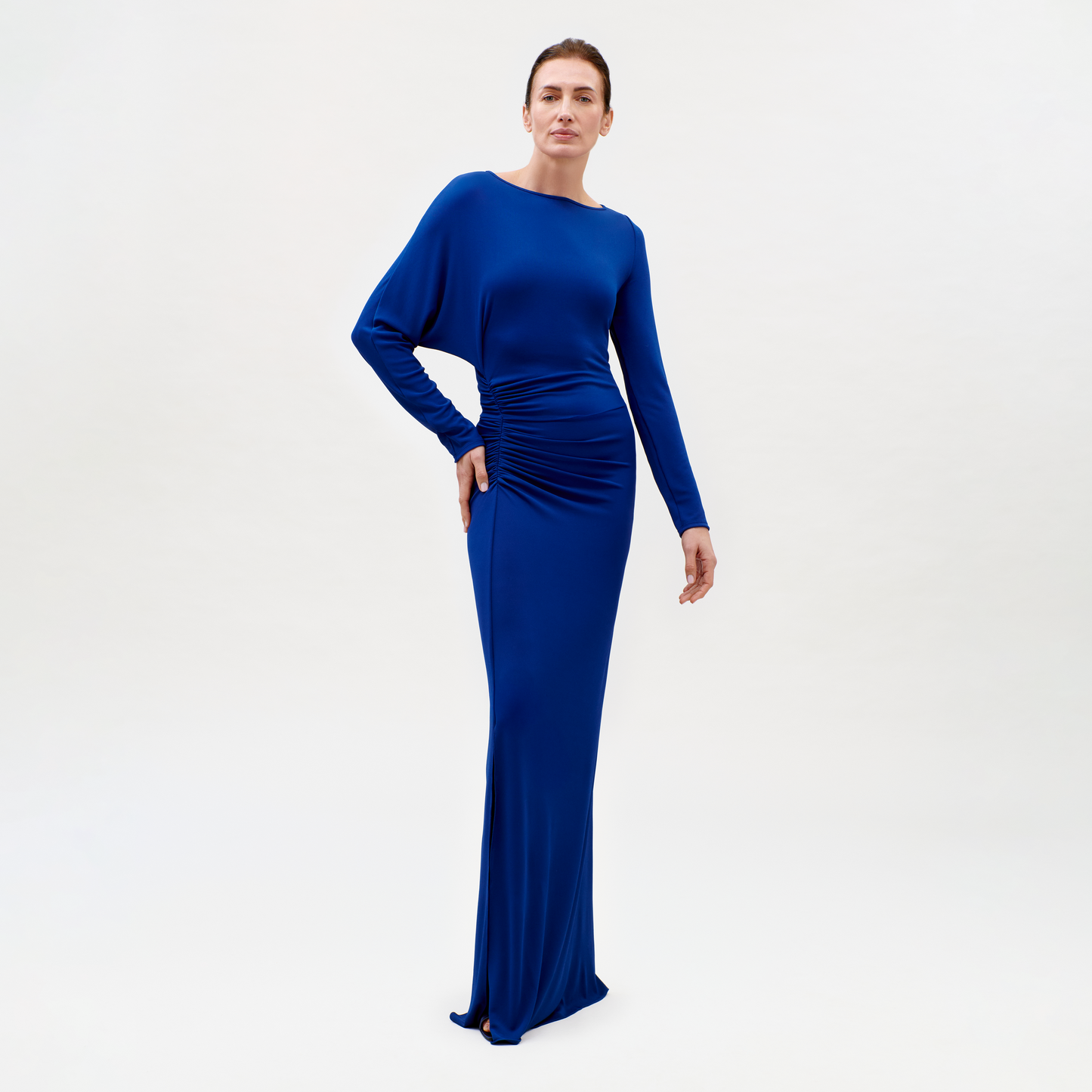 A woman stands against a plain background wearing the Laken Dress—a long, fitted, royal blue design with long sleeves and ruched waist detail. She poses with one hand on her hip, looking confidently at the camera.