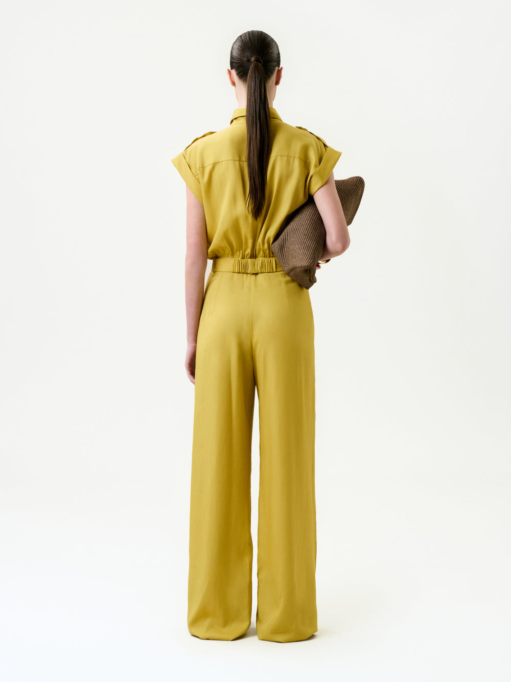 A person with long dark hair in a low ponytail wears the Sabrina Jumpsuit—a mustard yellow, short-sleeve, wide-leg lyocell piece—while holding a brown textured clutch under one arm against a plain white background.