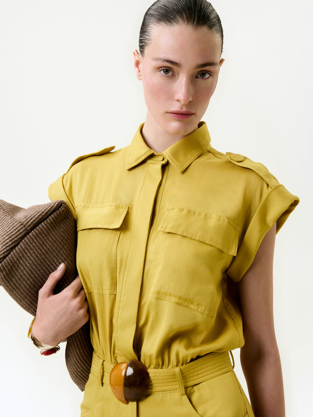 A woman with slicked-back hair and a neutral expression stands against a plain light background, wearing the Sabrina Jumpsuit—a mustard yellow, short-sleeve style in soft Italian fabric with chest pockets and a wide belt—holding a brown hat under her arm.