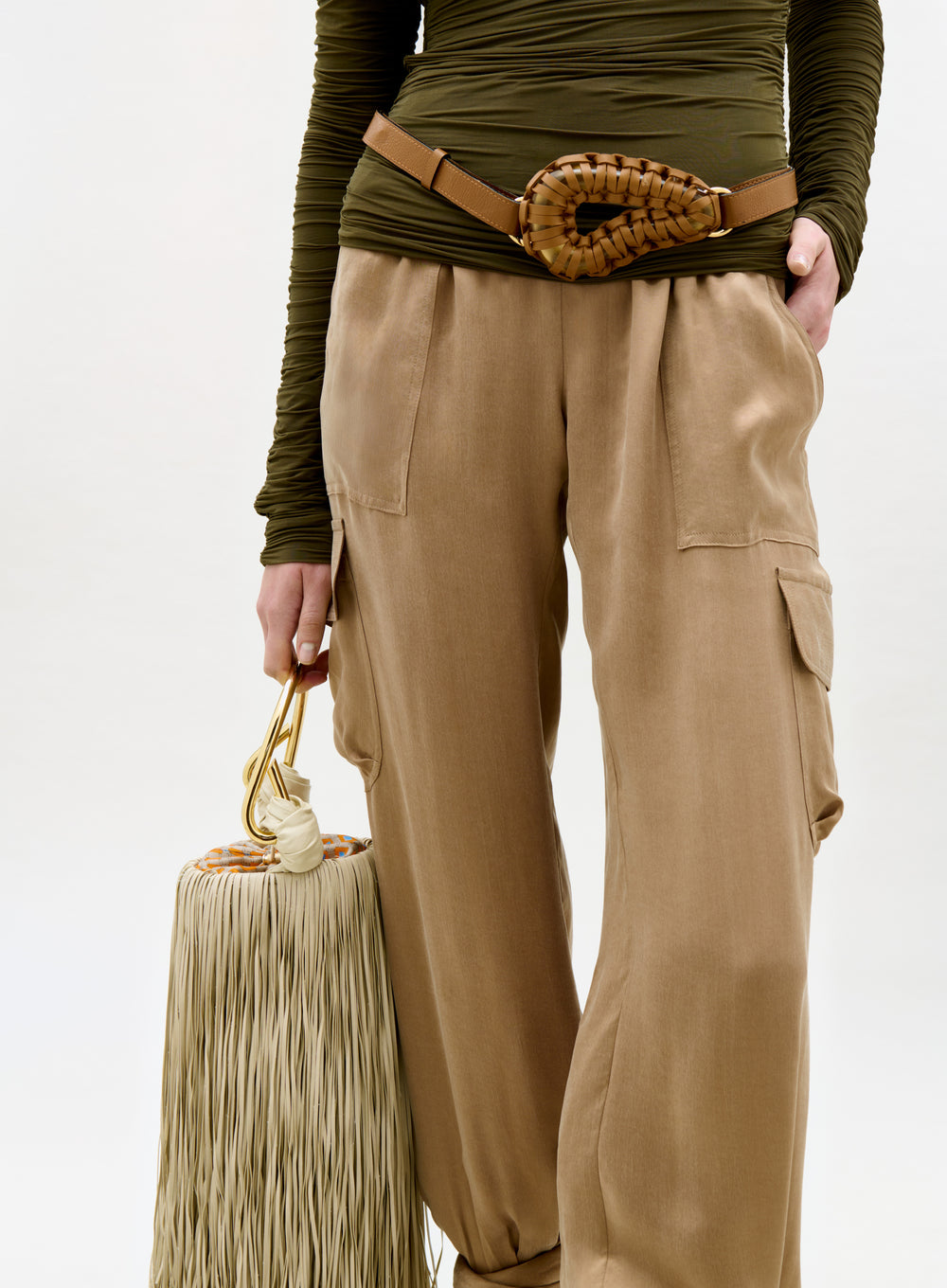 Person wearing a green top and beige cargo pants holding a beige handbag on a white background