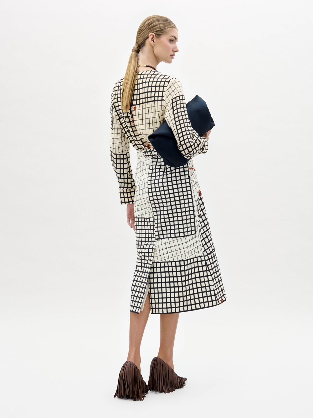 A woman stands against a white background wearing the Kadira Dress, a checked dress with red leaf prints, paired with a black clutch and brown fringed shoes.