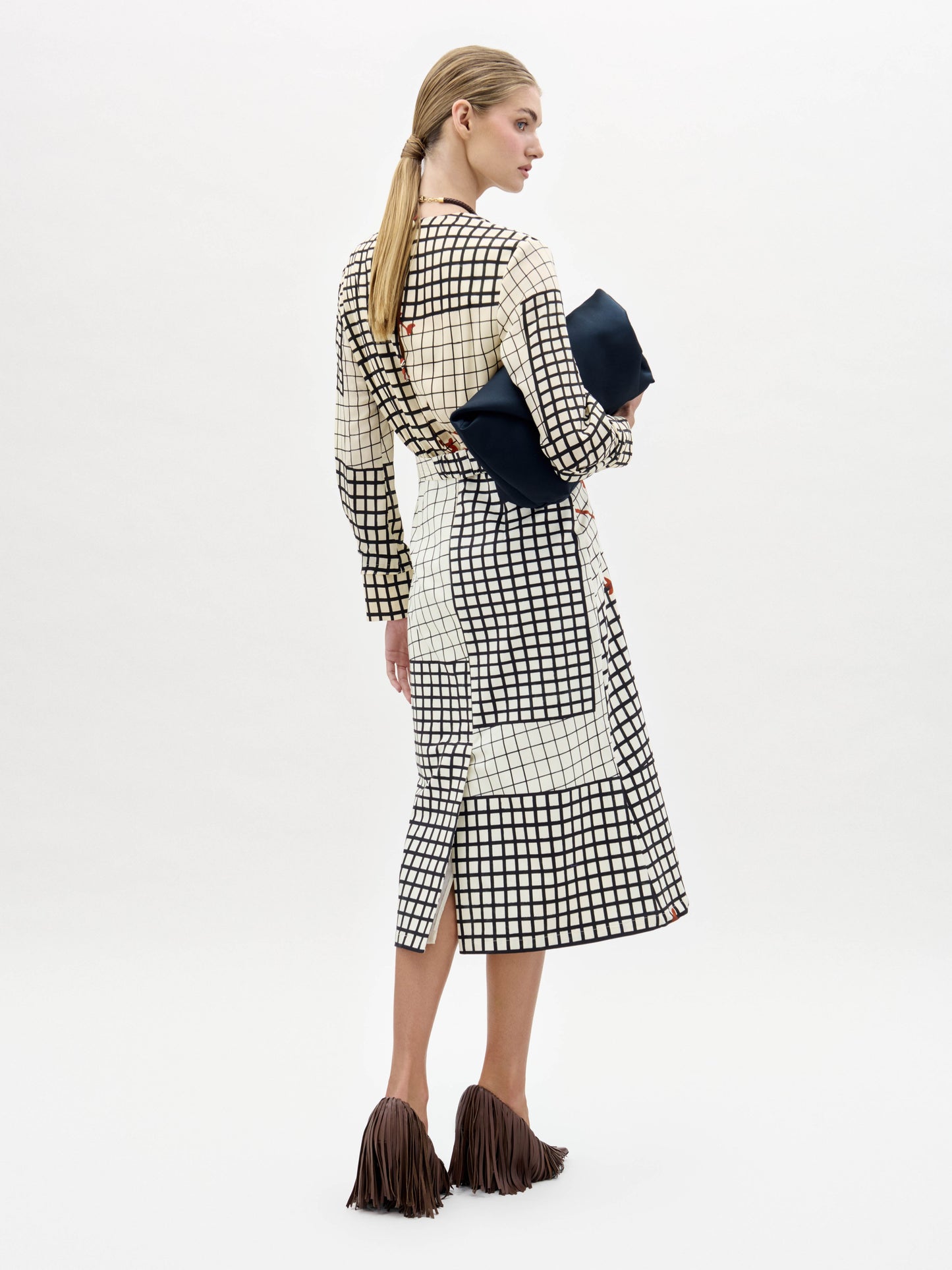 A woman stands against a white background wearing the Kadira Dress, a checked dress with red leaf prints, paired with a black clutch and brown fringed shoes.