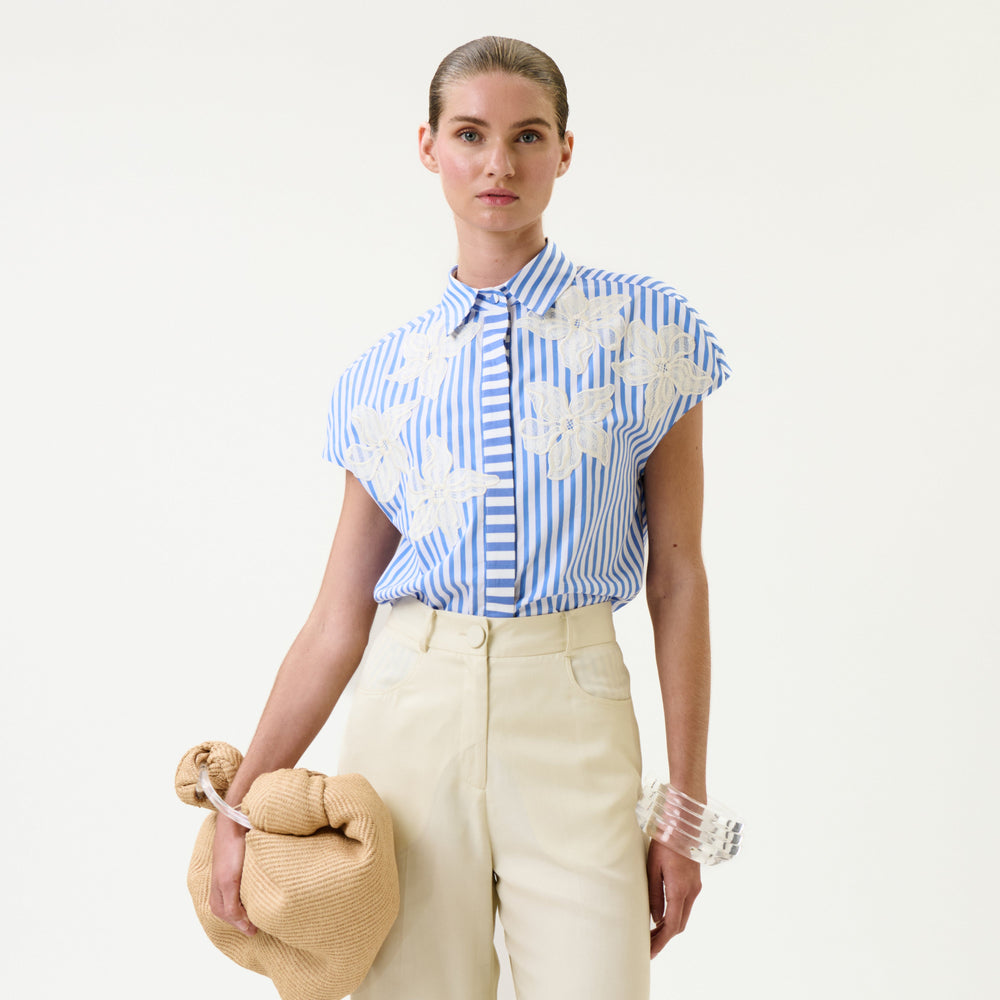 A woman in high-waisted cream pants wears the Maribel Shirt—a blue-and-white striped blouse with white floral patterns—paired with a tan knotted bag and a clear bangle, standing against a plain white background.