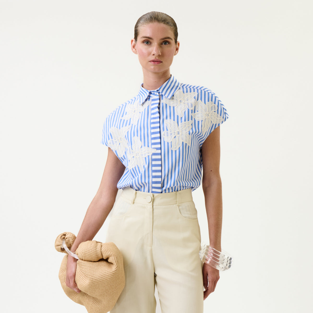 A woman in high-waisted cream pants wears the Maribel Shirt—a blue-and-white striped blouse with white floral patterns—paired with a tan knotted bag and a clear bangle, standing against a plain white background.
