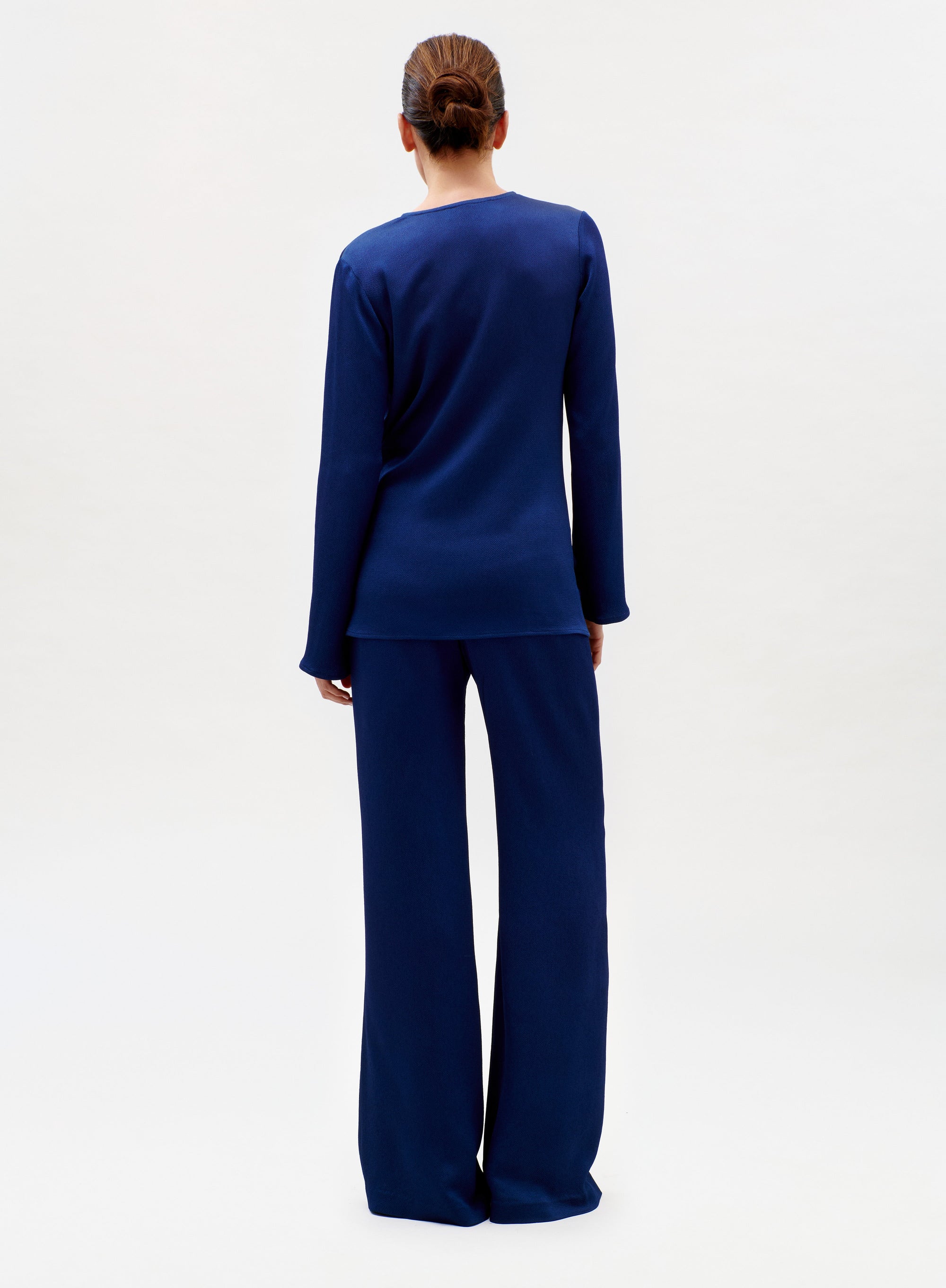 A person with brown hair in a low bun stands facing away, wearing a dark blue long-sleeve top and the matching Palermo Pant in wide-leg style against a plain white background.
