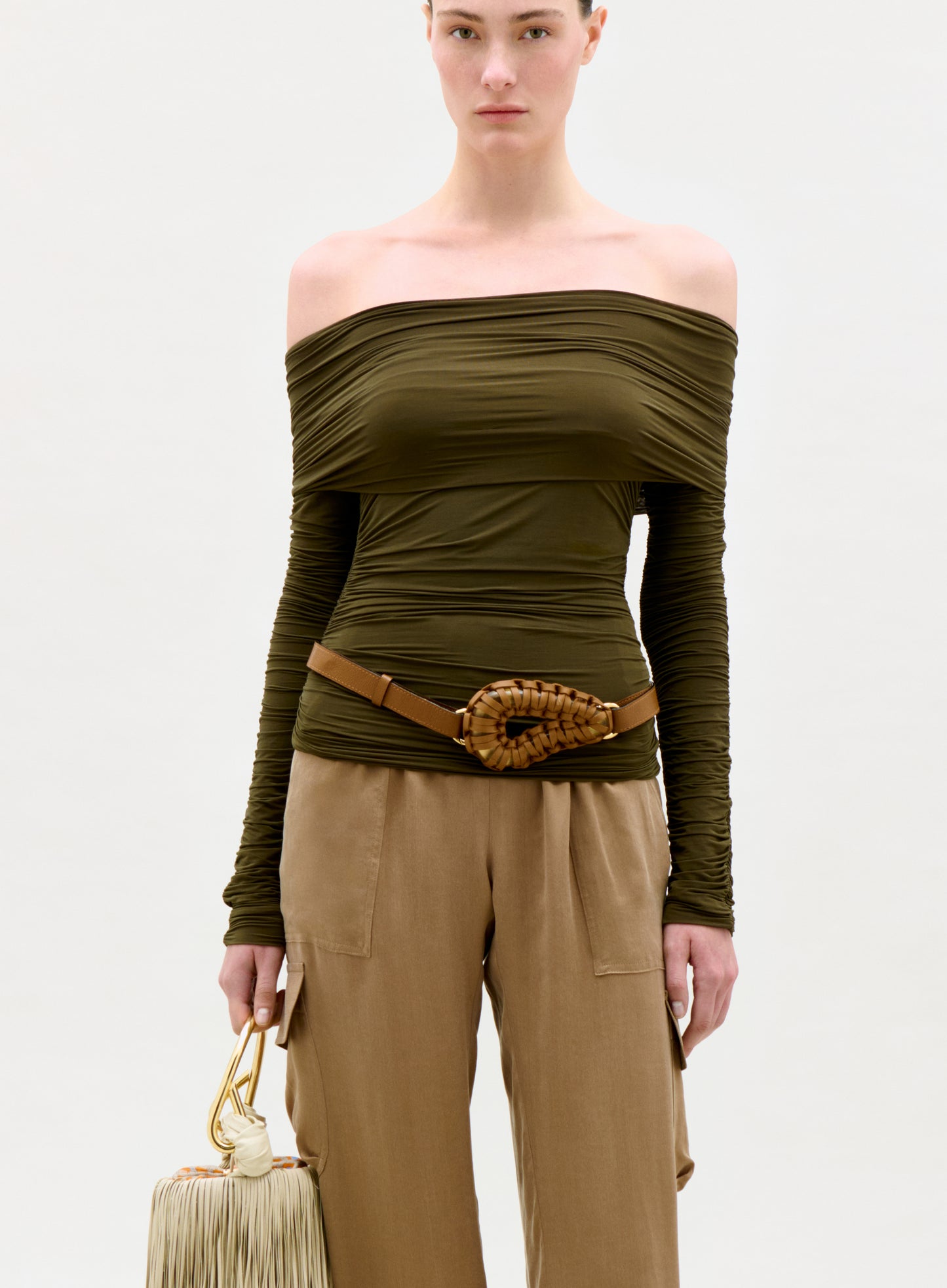 Woman wearing an olive green off-shoulder top and beige pants, holding a beige handbag on a white background.