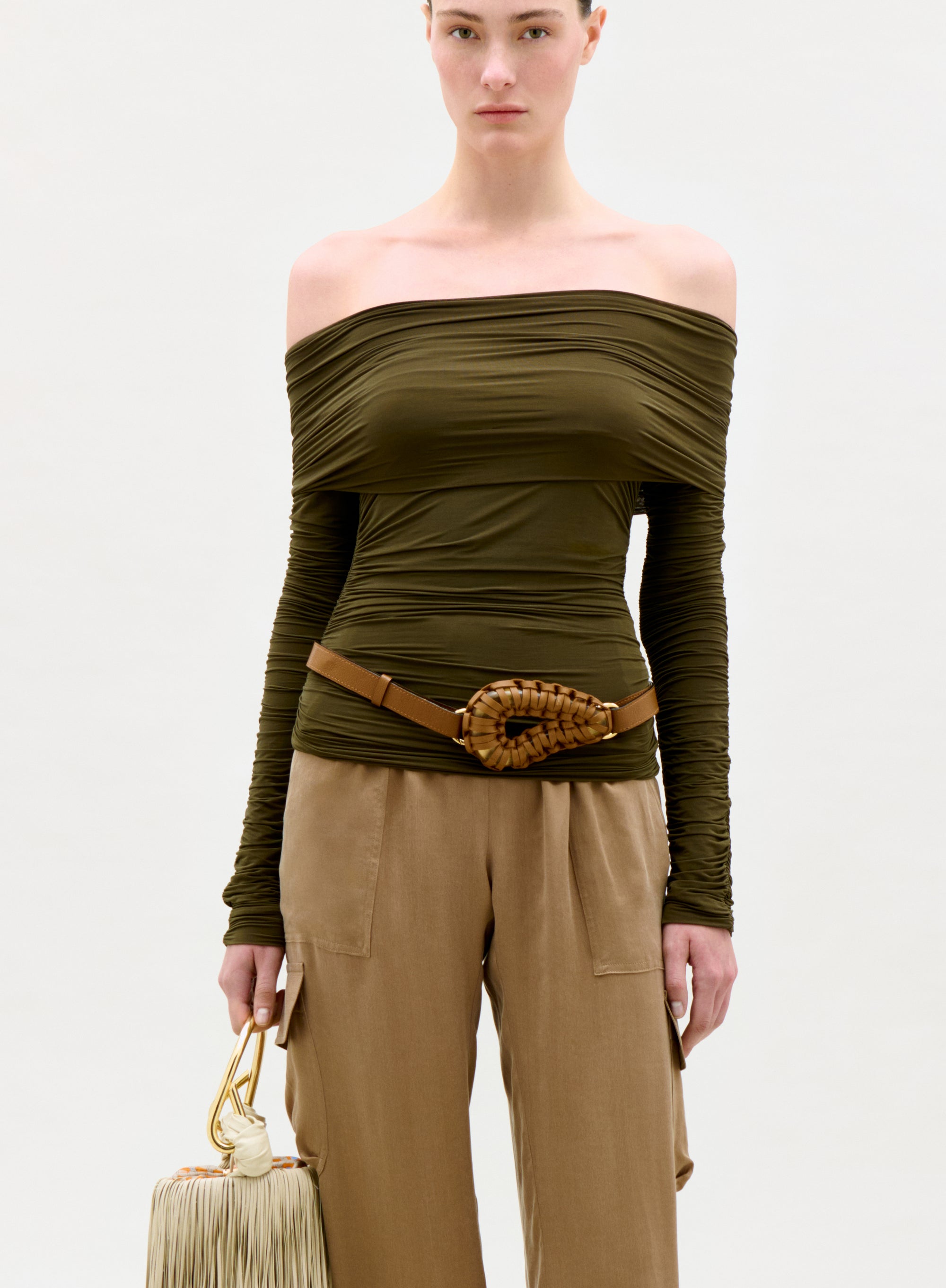 Woman wearing an olive green off-shoulder top and beige pants, holding a beige handbag on a white background.