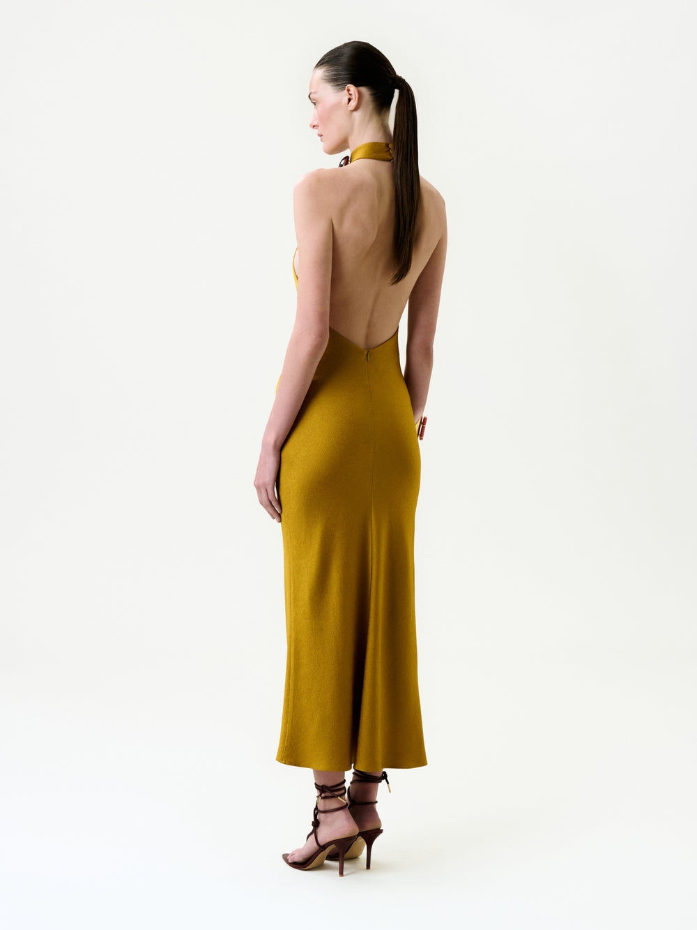 A woman with long dark hair in a ponytail stands in profile against a plain white background, wearing the mustard yellow backless halter Lali Dress crafted from Italian fabric and strappy brown high heels.
