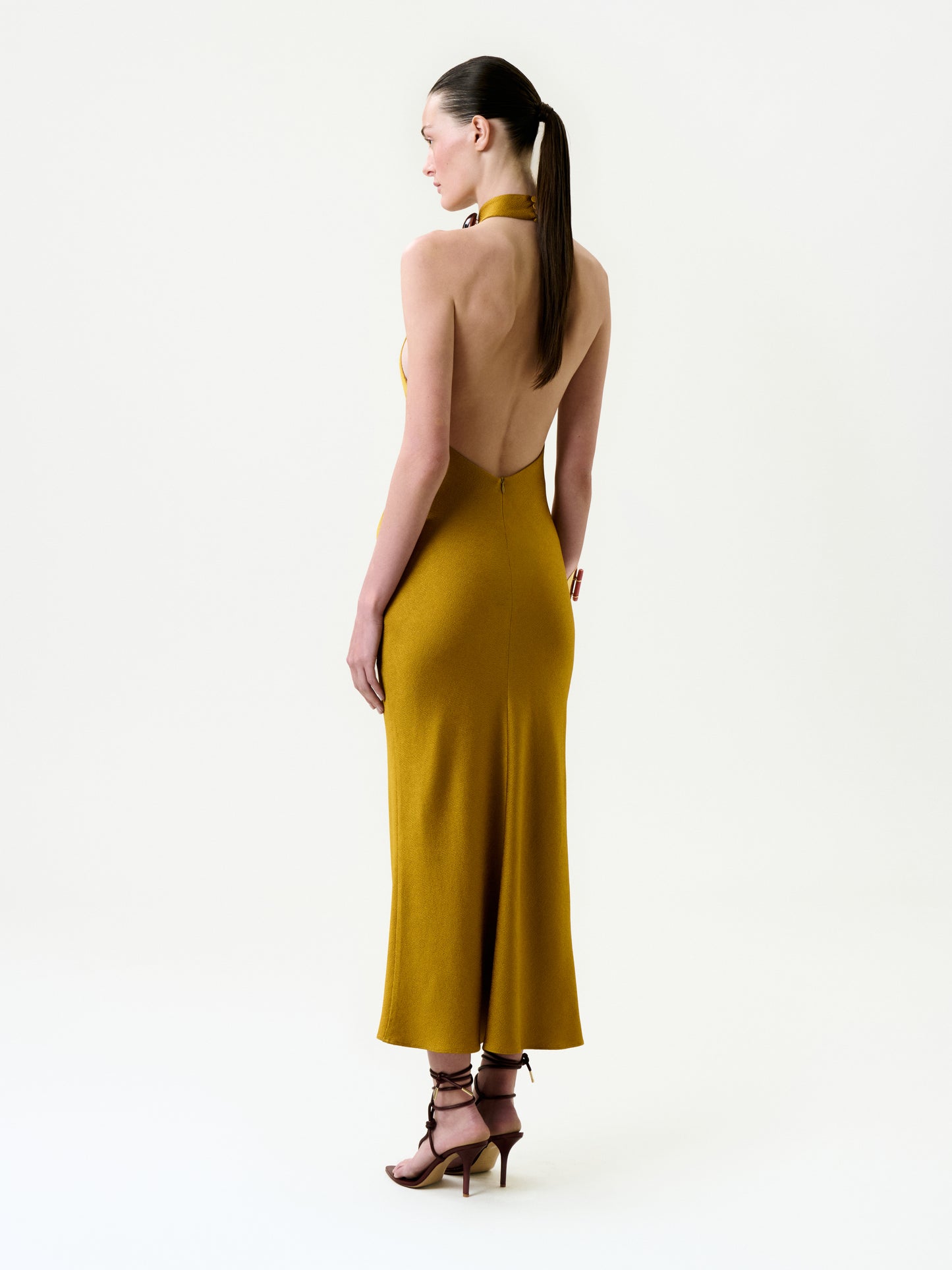 A woman with long dark hair in a ponytail stands in profile against a plain white background, wearing the mustard yellow backless halter Lali Dress crafted from Italian fabric and strappy brown high heels.