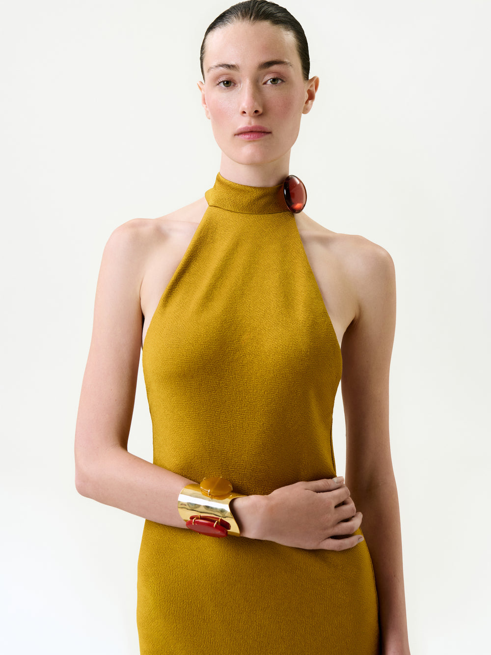 A woman with slicked-back hair models the Lali Dress—a mustard yellow halter-neck midi made in Portugal from fine Italian fabric—paired with chunky gold and red bracelets, set against a plain light background.