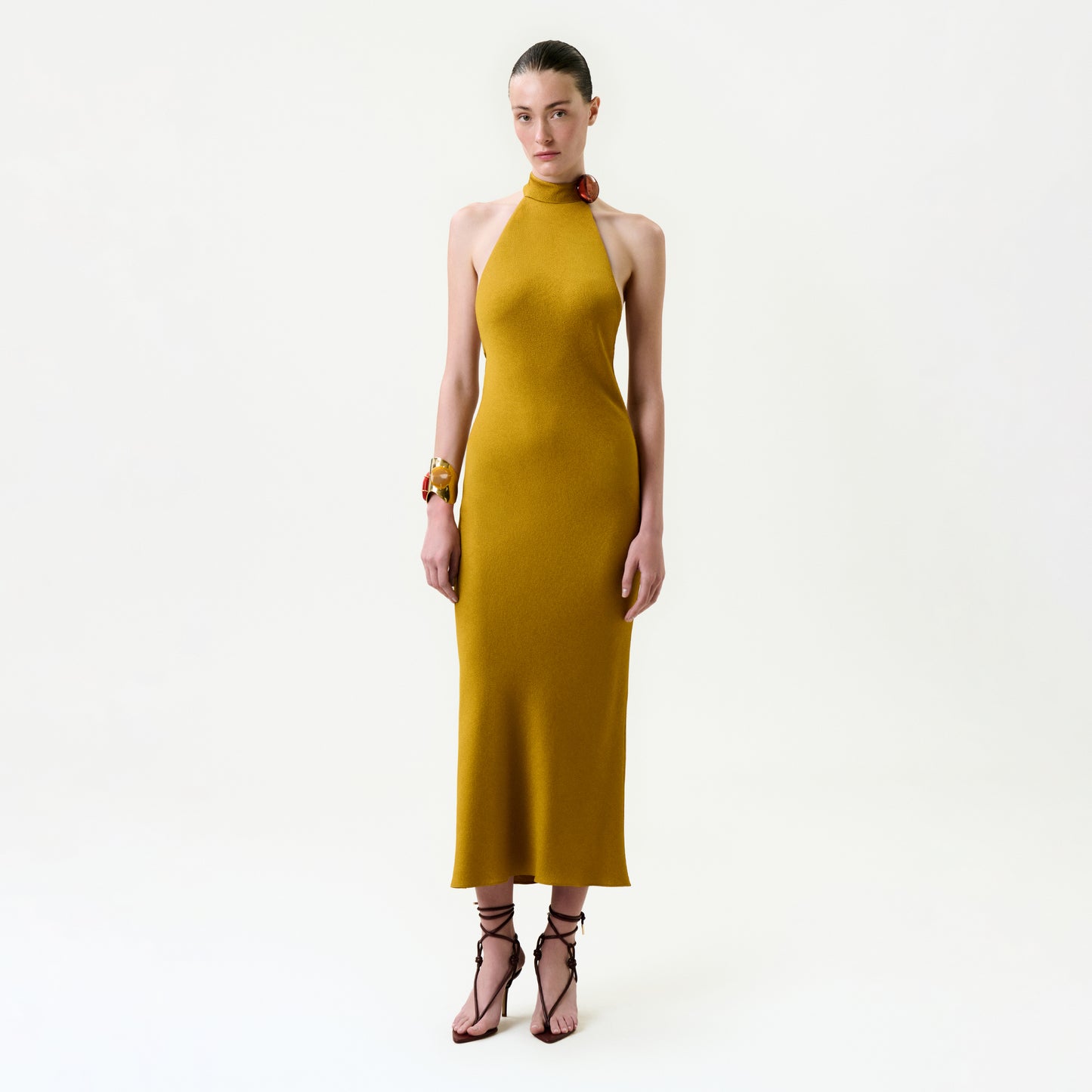 A woman with slicked-back hair and a neutral expression wears the Lali Dress—an Italian fabric mustard yellow midi—paired with bracelets and strappy high-heeled sandals, standing against a plain white background.
