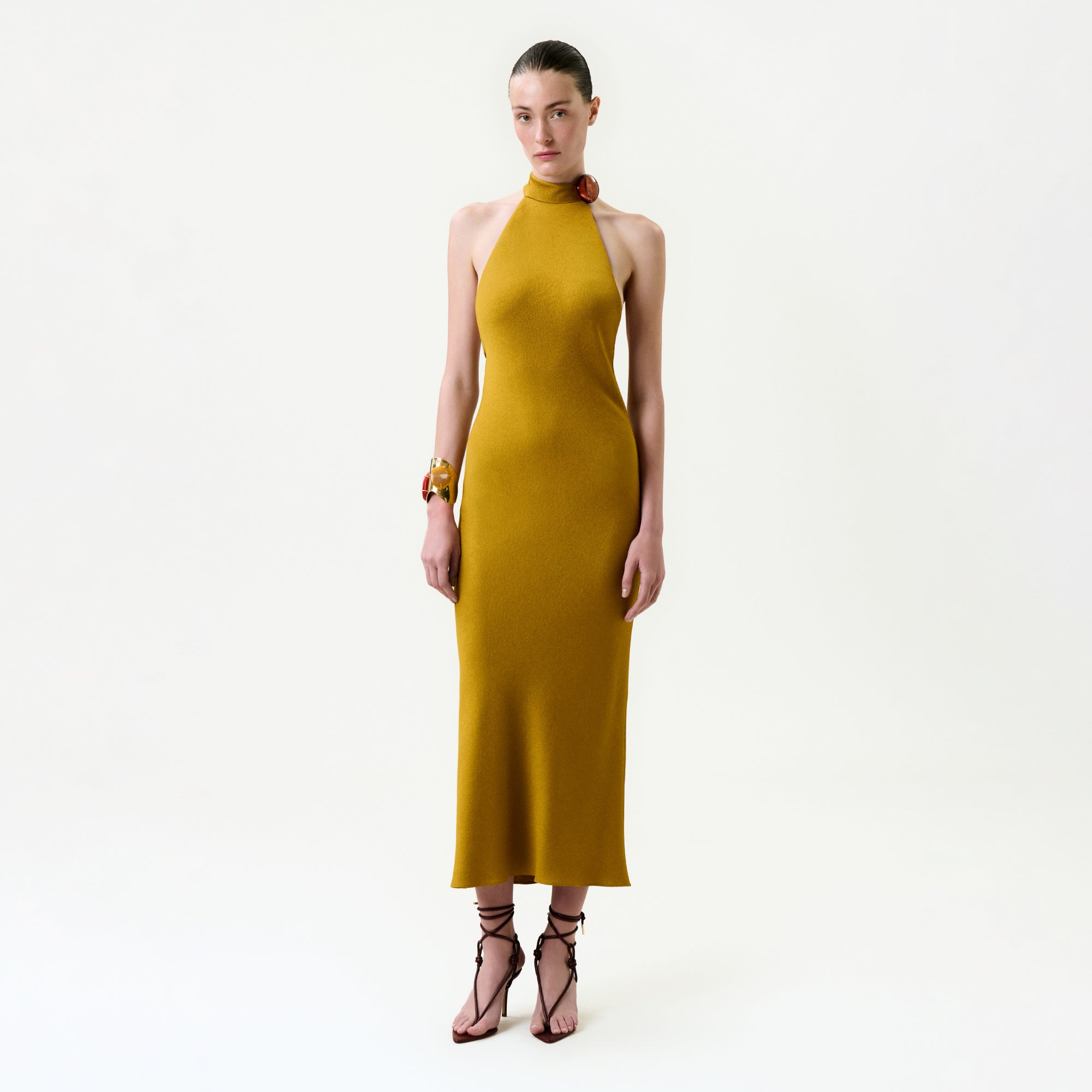 A woman with slicked-back hair and a neutral expression wears the Lali Dress—an Italian fabric mustard yellow midi—paired with bracelets and strappy high-heeled sandals, standing against a plain white background.