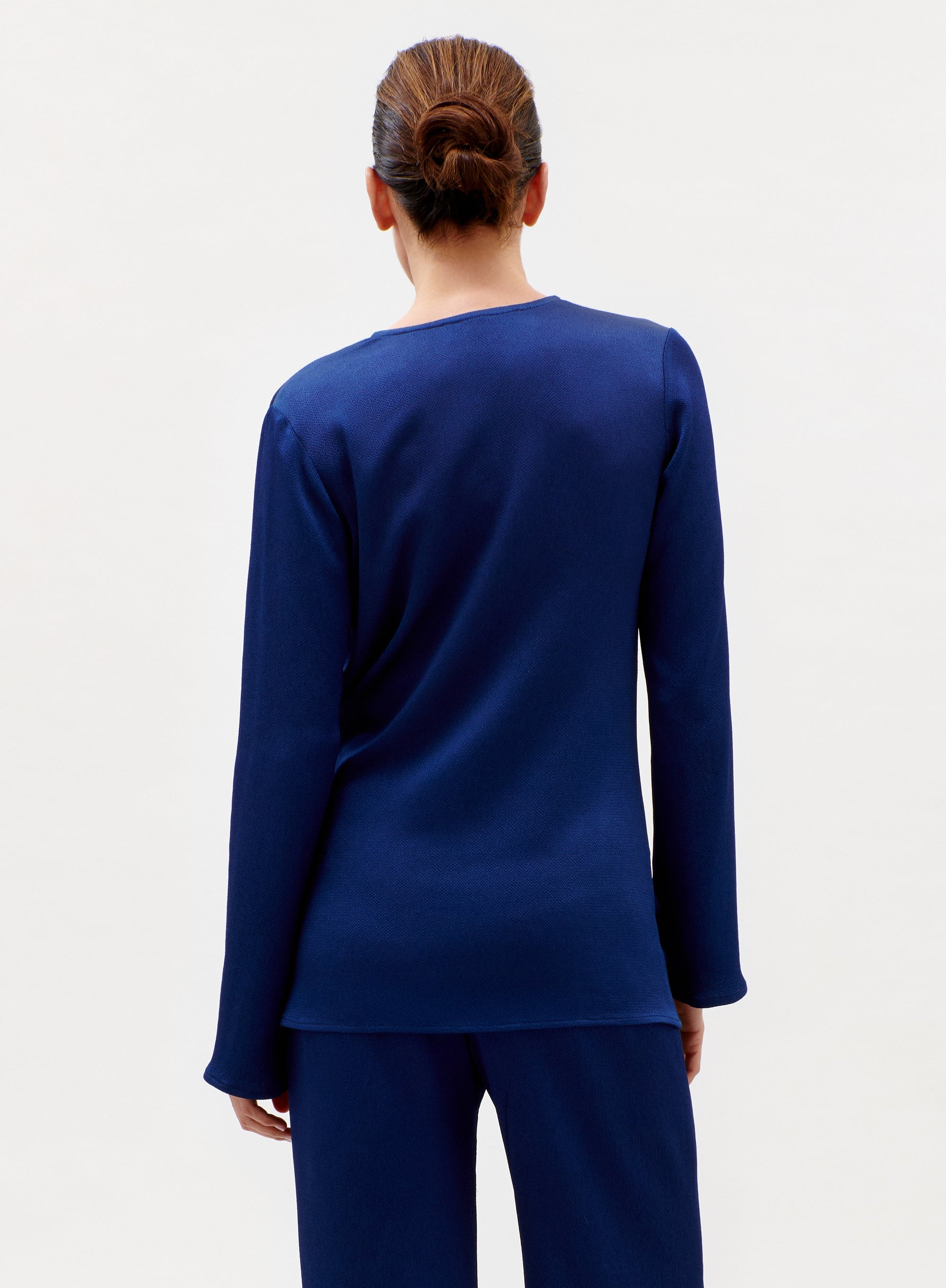 A woman with brown hair in a low bun stands with her back to the camera, wearing the long-sleeved, dark blue Tayra Blouse and matching pants against a plain light background.