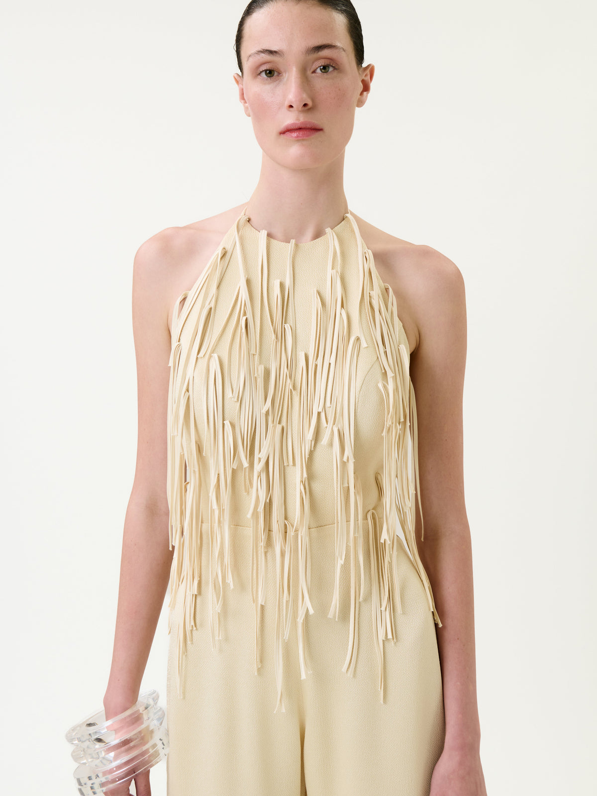 A person stands against a plain white background wearing the Tulsi Jumpsuit, a beige sleeveless piece featuring long fringe details on the front, and holds a chunky clear bracelet.