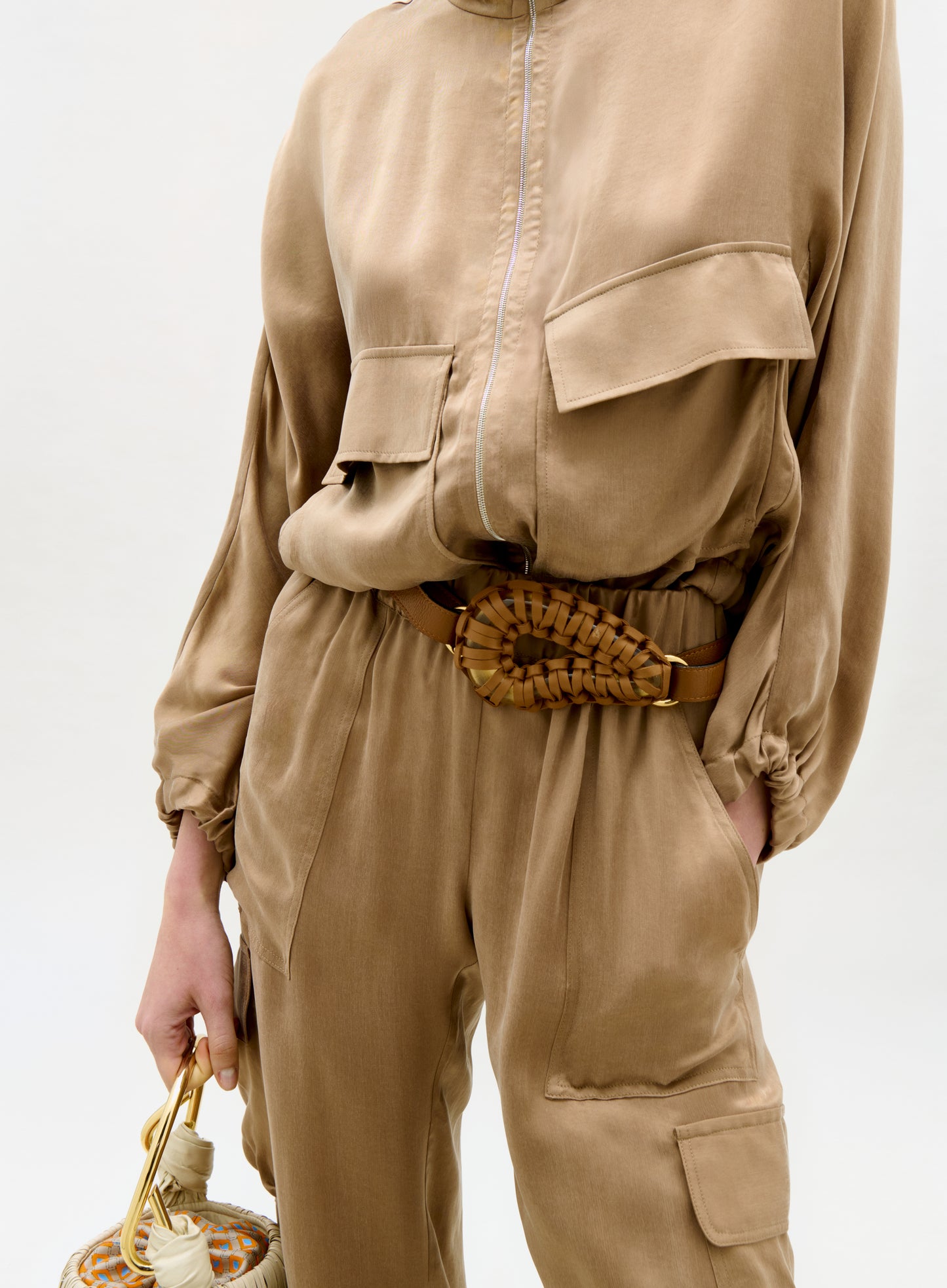 Person wearing a beige jumpsuit with a belt and holding a handbag on a white background