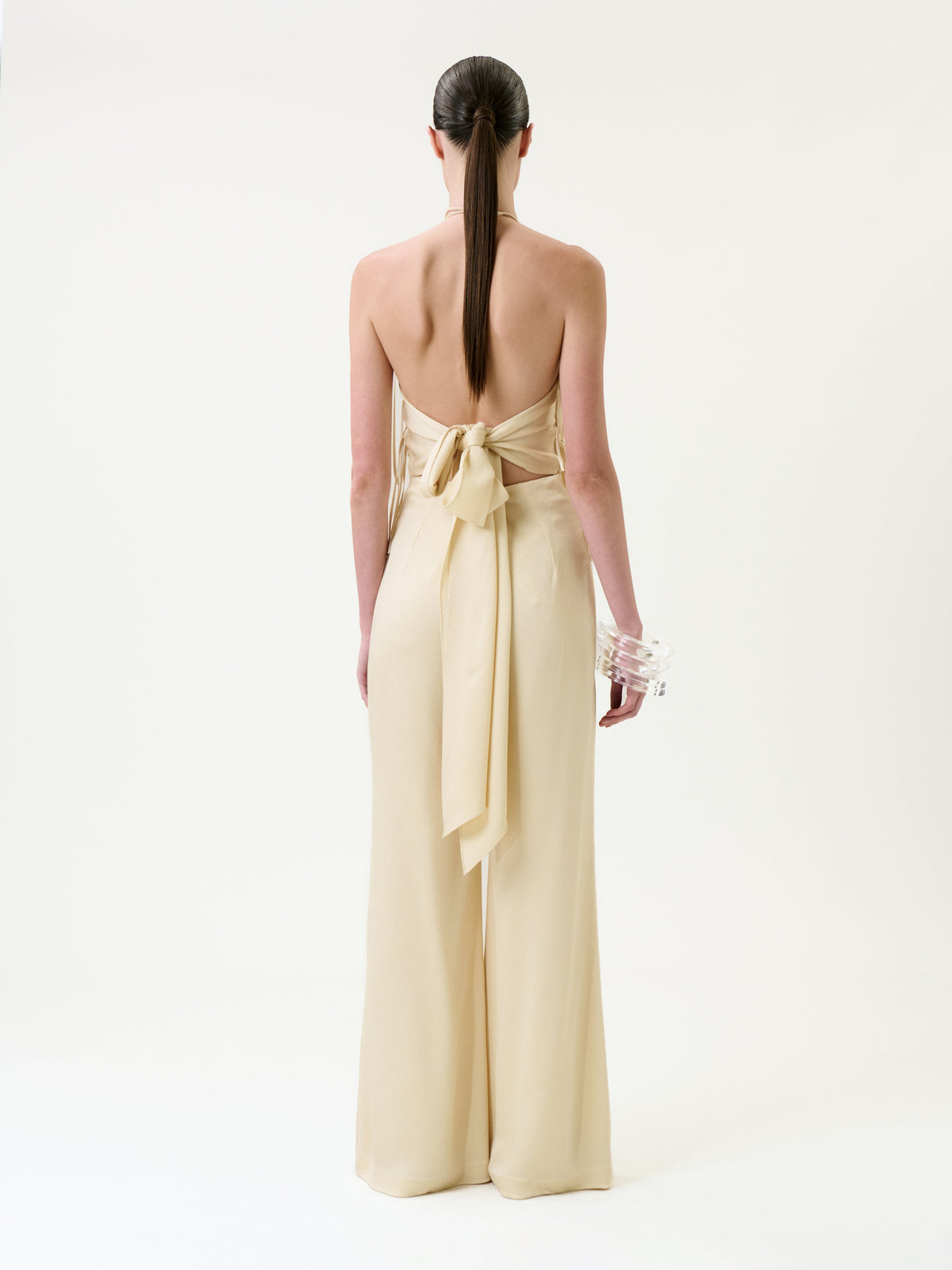 Woman with long dark hair in a low ponytail, wearing the backless Tulsi Jumpsuit in beige, featuring a large bow at the waist, stands facing away on a white background, holding clear bangles in one hand.