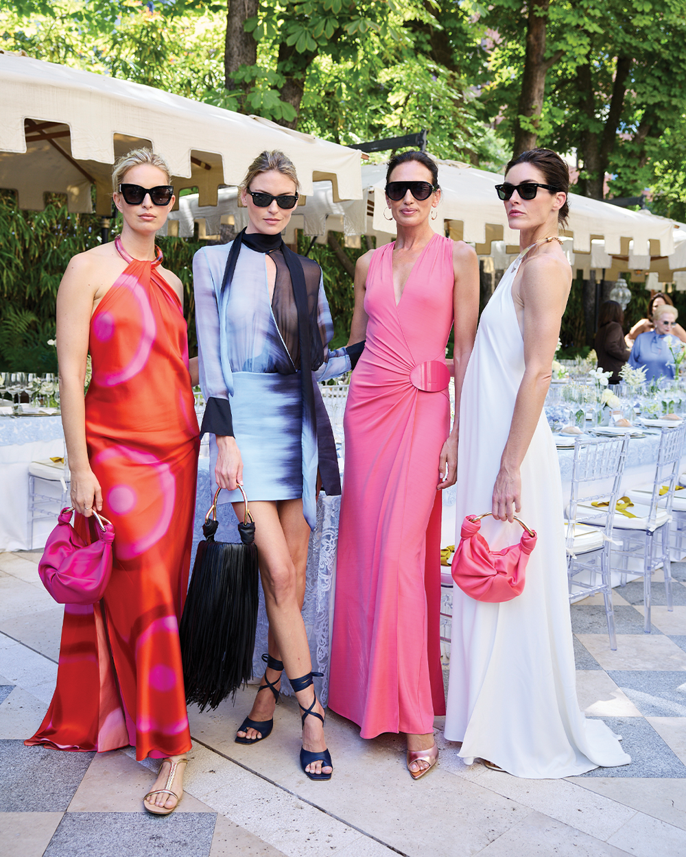 Five women in stylish outfits standing outdoors with tables and chairs in the background.