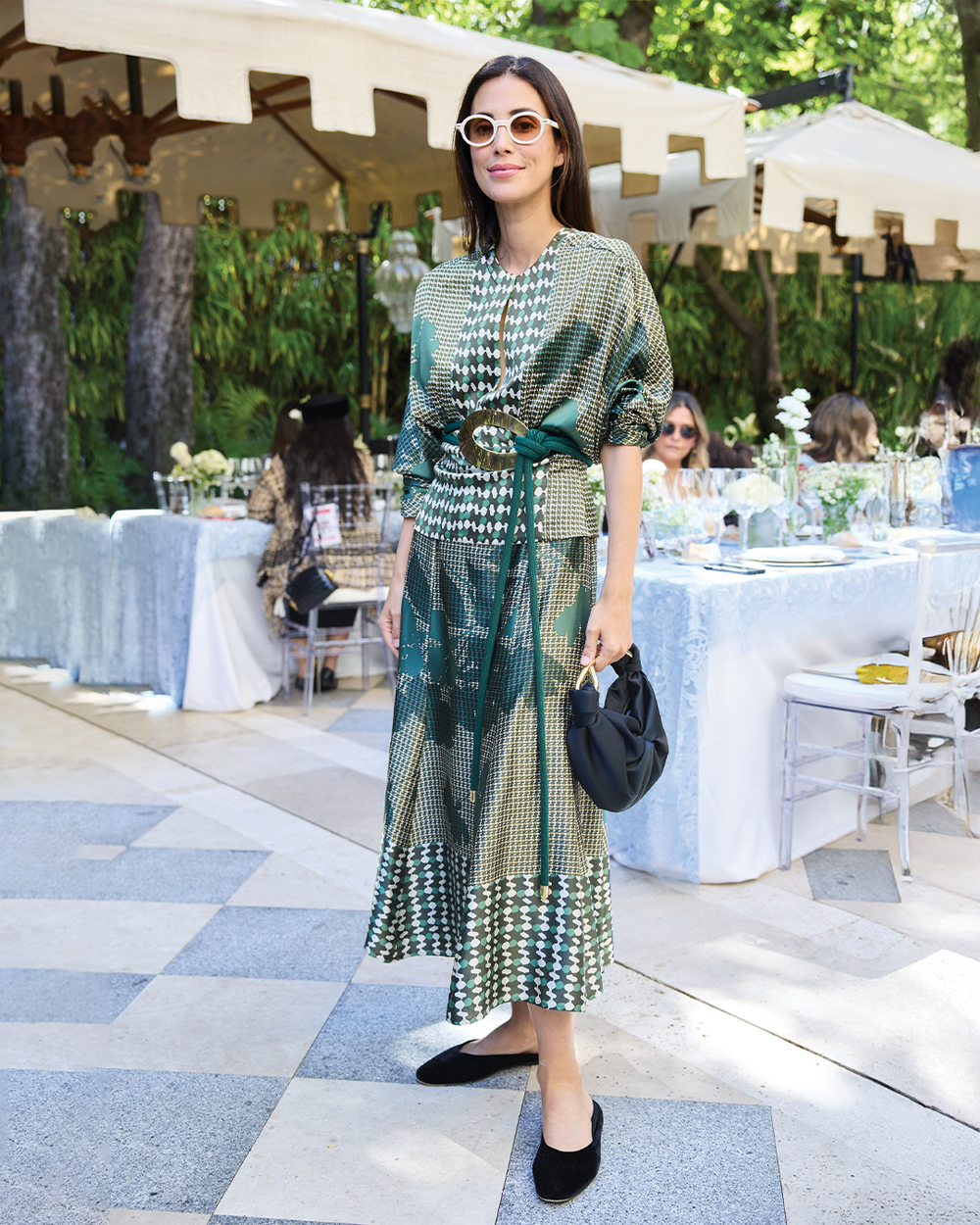 Woman in a green patterned dress standing outdoors at an event with tables and umbrellas.