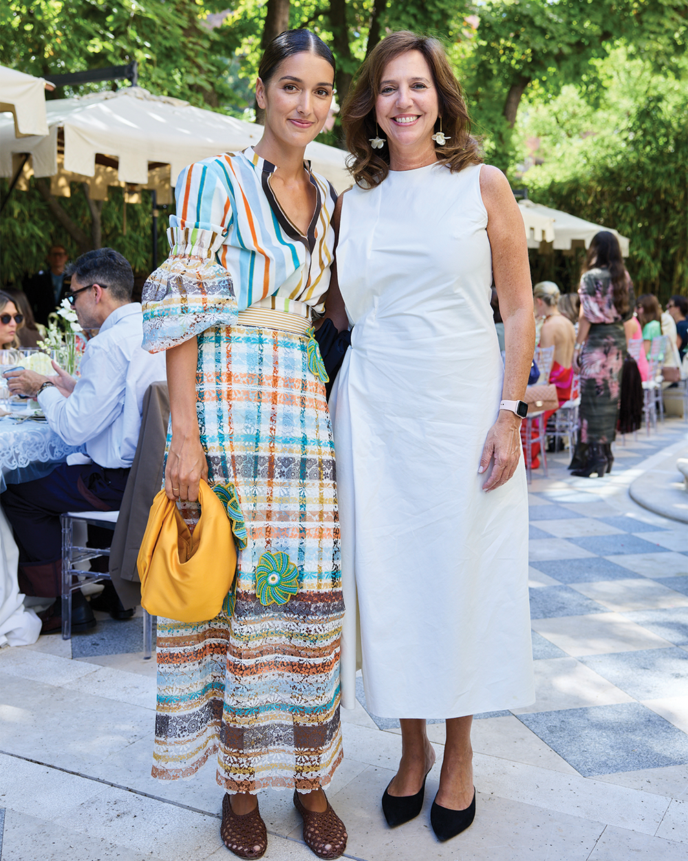 Two women standing outdoors in a social setting, one in a colorful dress and the other in a white dress.
