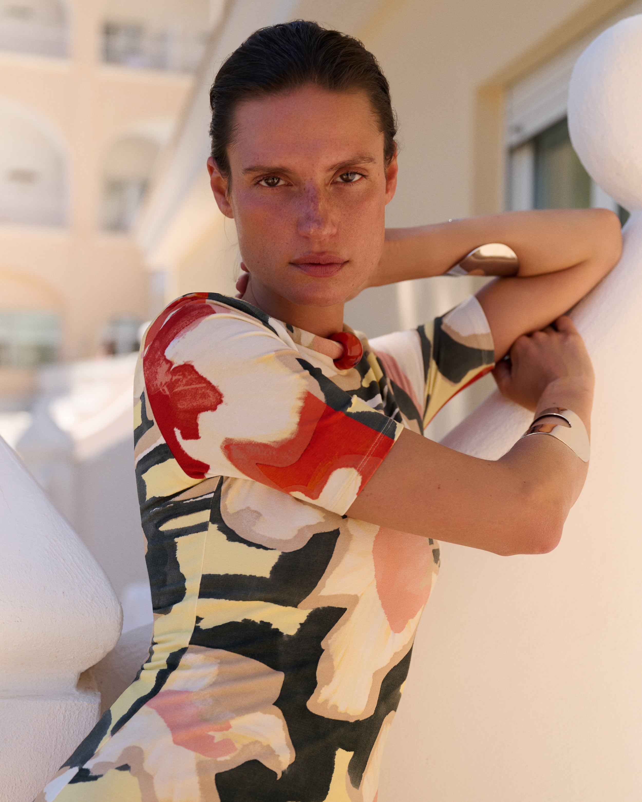 A woman wearing a short-sleeve abstract print top with red, black, cream, and pink brushstroke patterns, leaning against a white architectural structure.