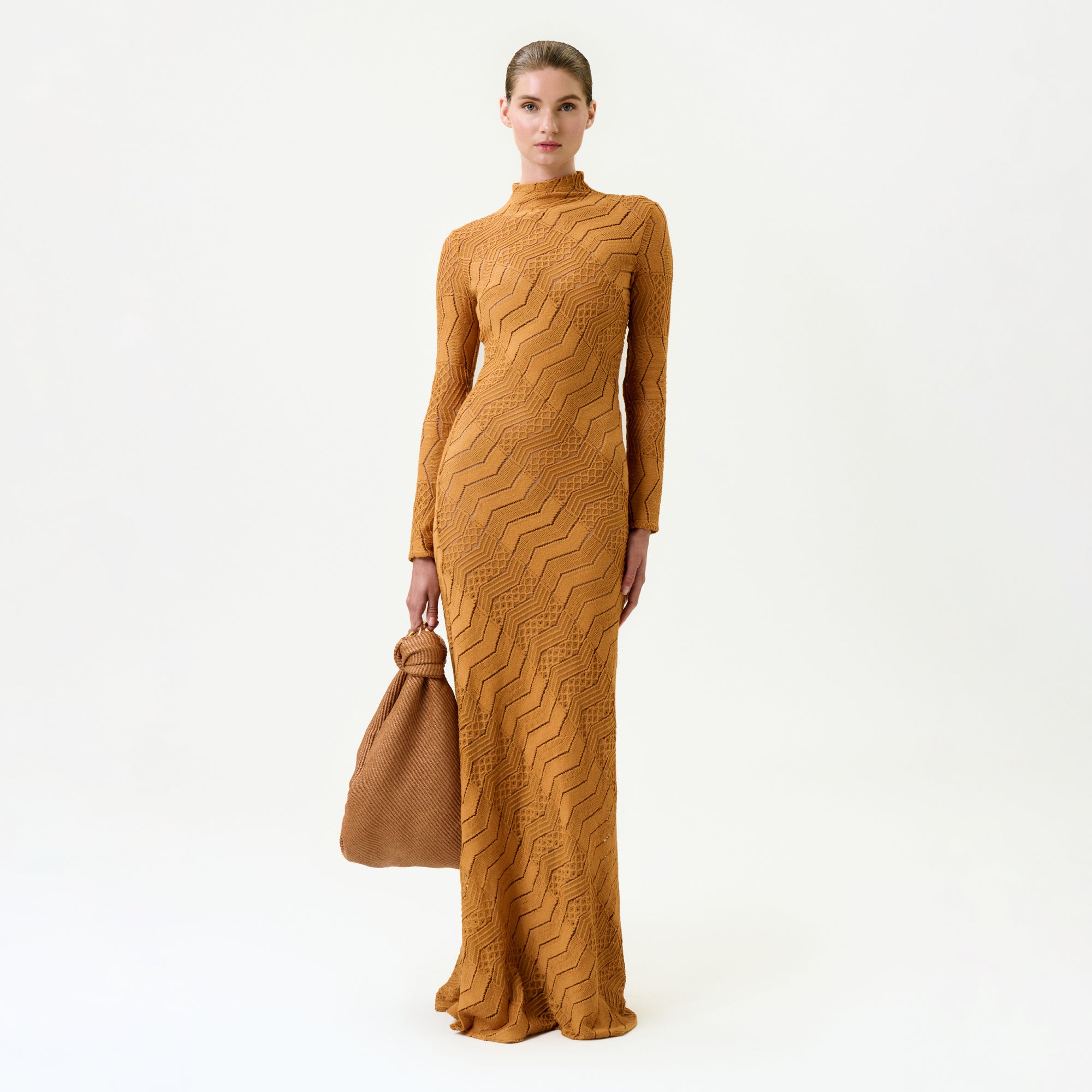 A woman in the Marianne Dress—an orange, floor-length, long-sleeved maxi made from Italian fabric—stands with a matching handbag against a white background.