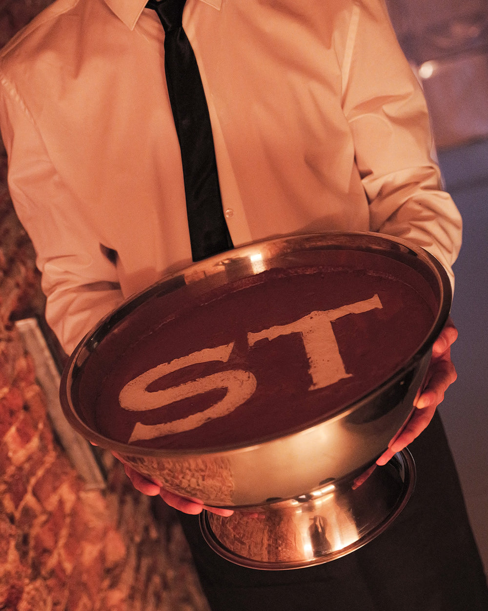 A person in a white dress shirt and black tie holds a large metal pedestal bowl filled with chocolate or cocoa, with the letters 'ST' molded into the surface.